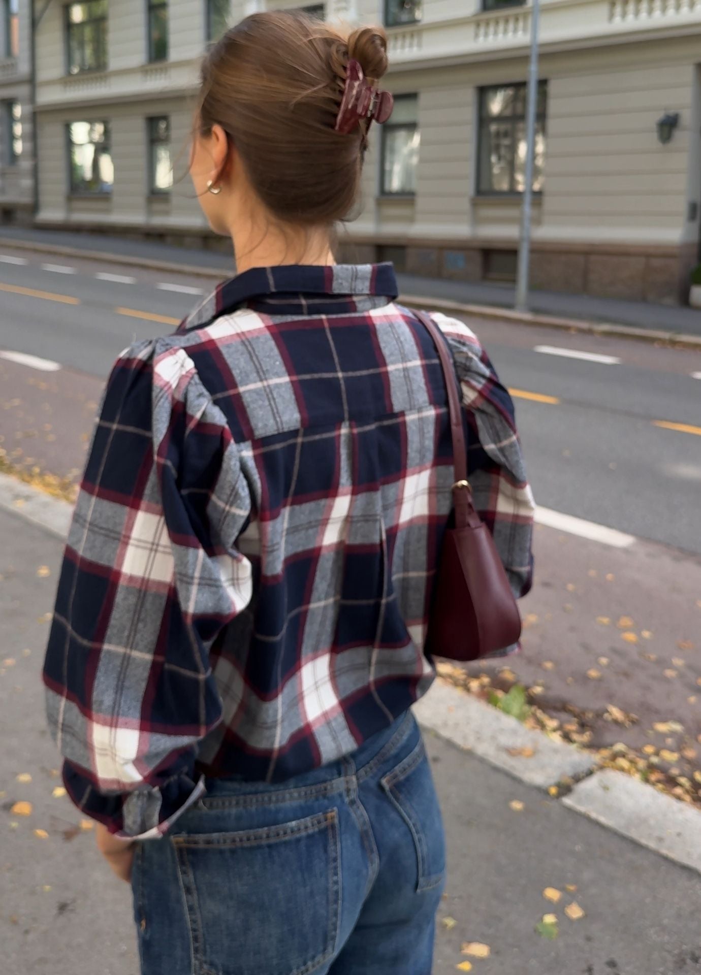 A woman viewed from behind wearing a navy, red, and white plaid flannel shirt with blue jeans, carrying a dark red shoulder bag on a city street.
