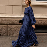 A woman wearing a long midnight blue satin gown with balloon sleeves and a tiered skirt walks along a city sidewalk next to a beige stone building.