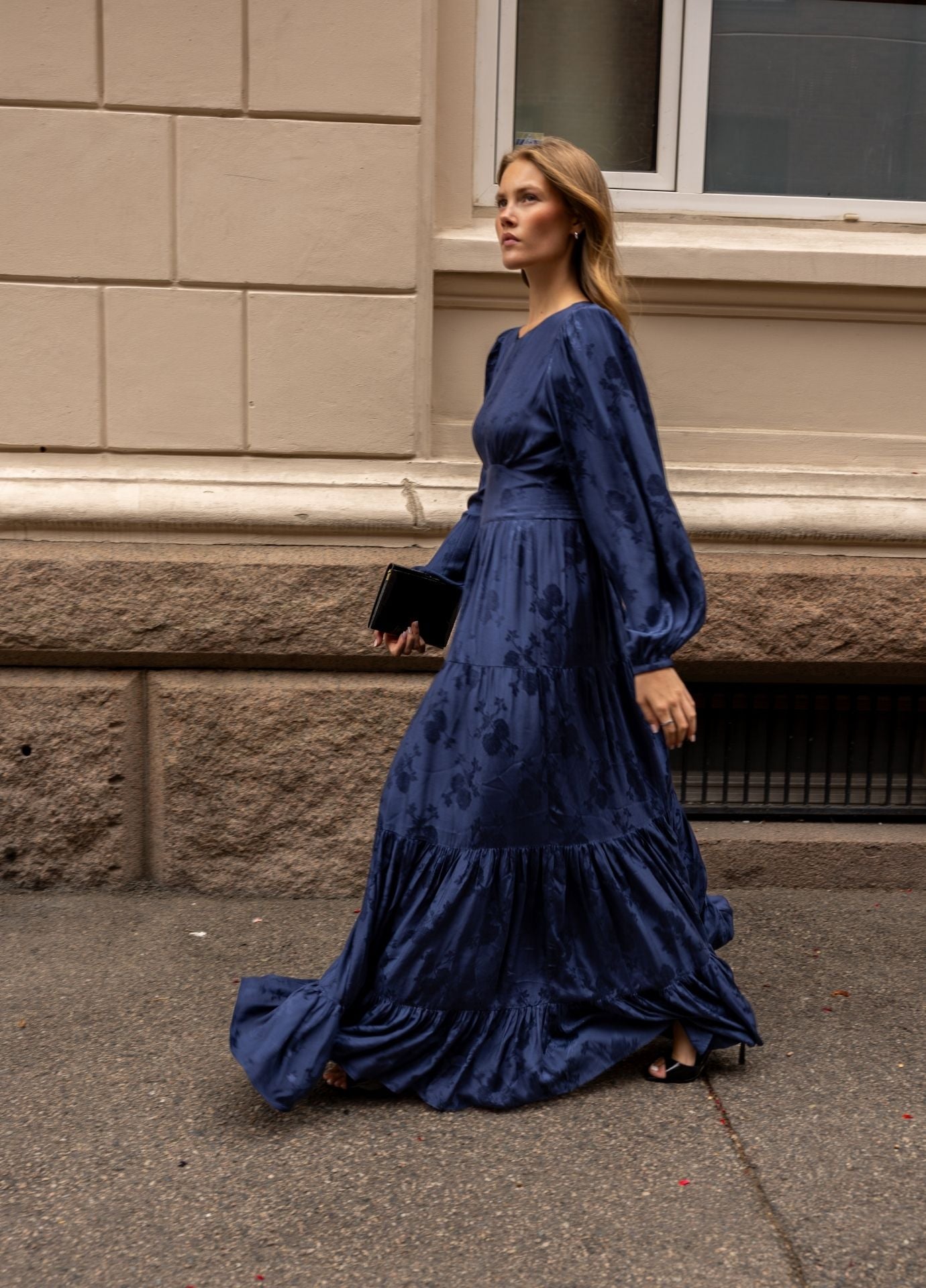 A woman wearing a long midnight blue satin gown with balloon sleeves and a tiered skirt walks along a city sidewalk next to a beige stone building.