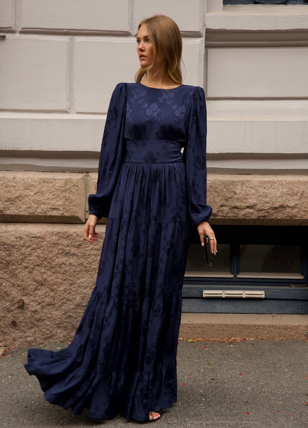 A woman wearing a long midnight blue maxi dress in viscose satin with a subtle tonal damask pattern, standing outdoors in front of a stone building wall.