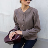 A woman wearing a taupe brown button-front cardigan, dark blue jeans, and black sunglasses, holding a burgundy leather handbag against a light gray concrete wall.