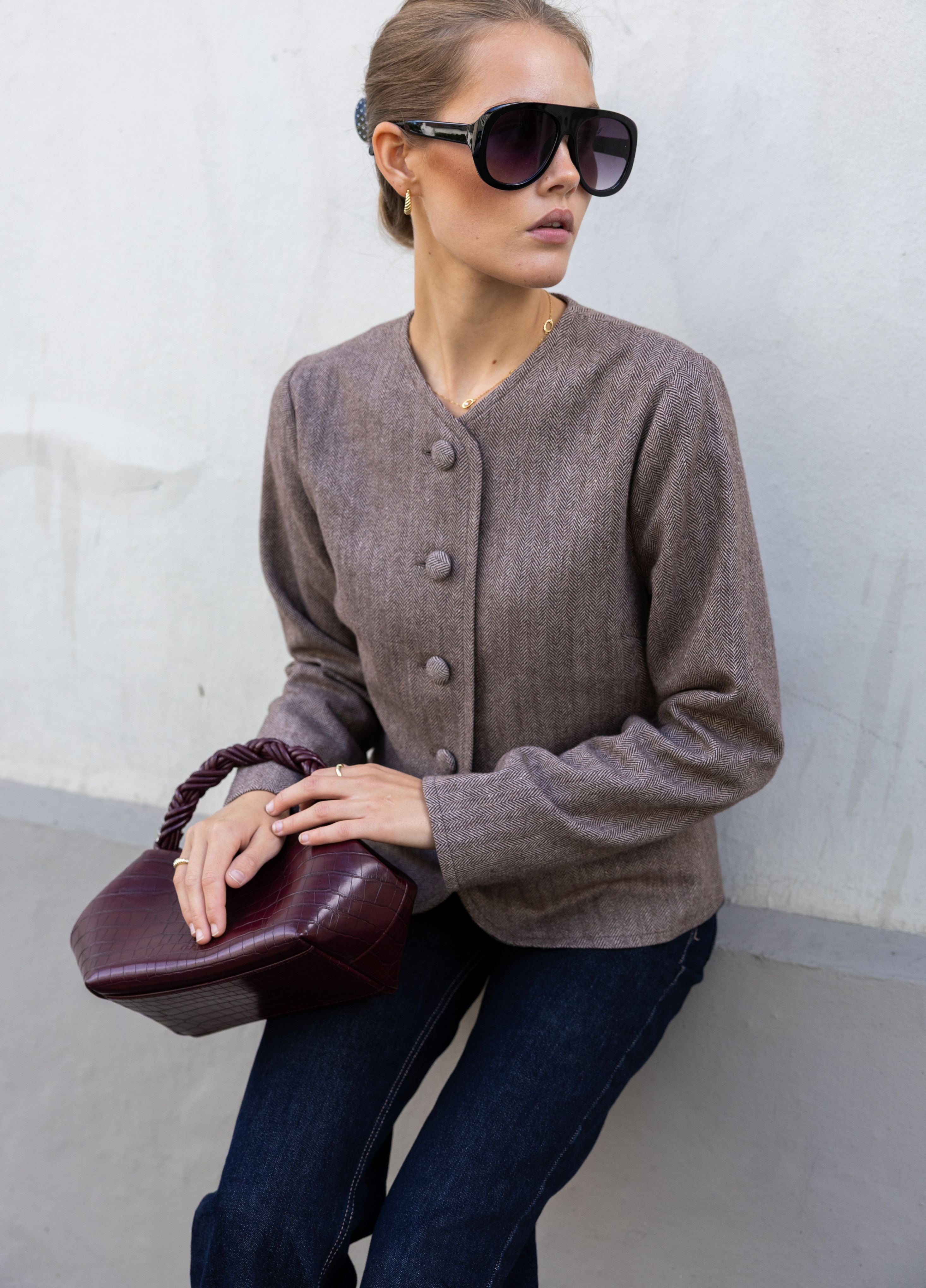 A woman wearing a taupe brown button-front cardigan, dark blue jeans, and black sunglasses, holding a burgundy leather handbag against a light gray concrete wall.