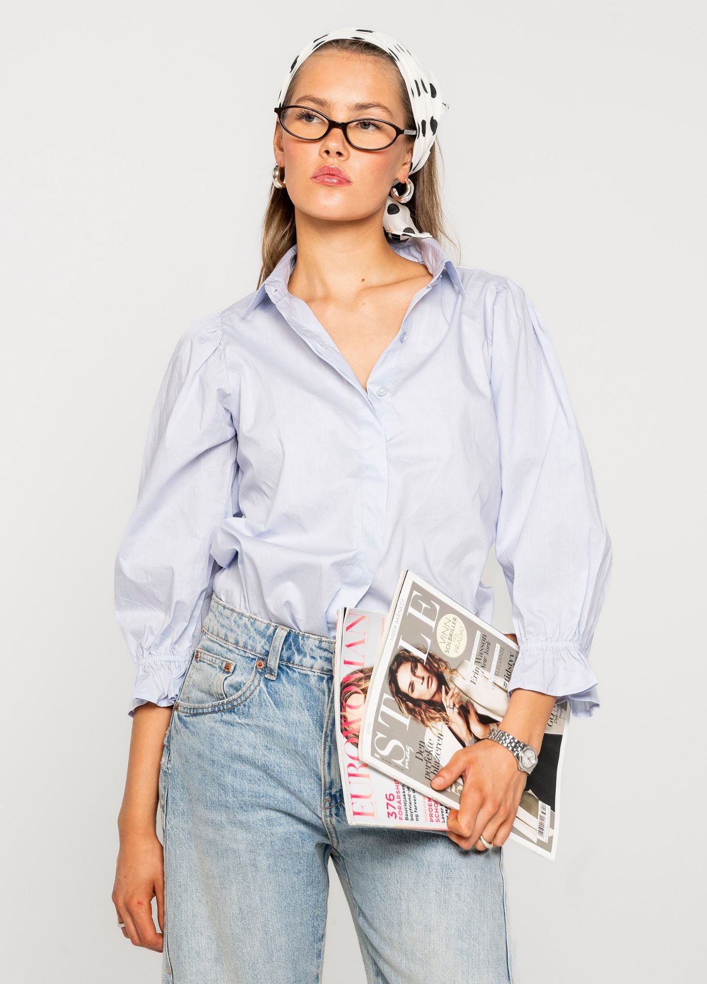 Woman wearing a blue shirt, jeans, and glasses holding a magazine against a white background