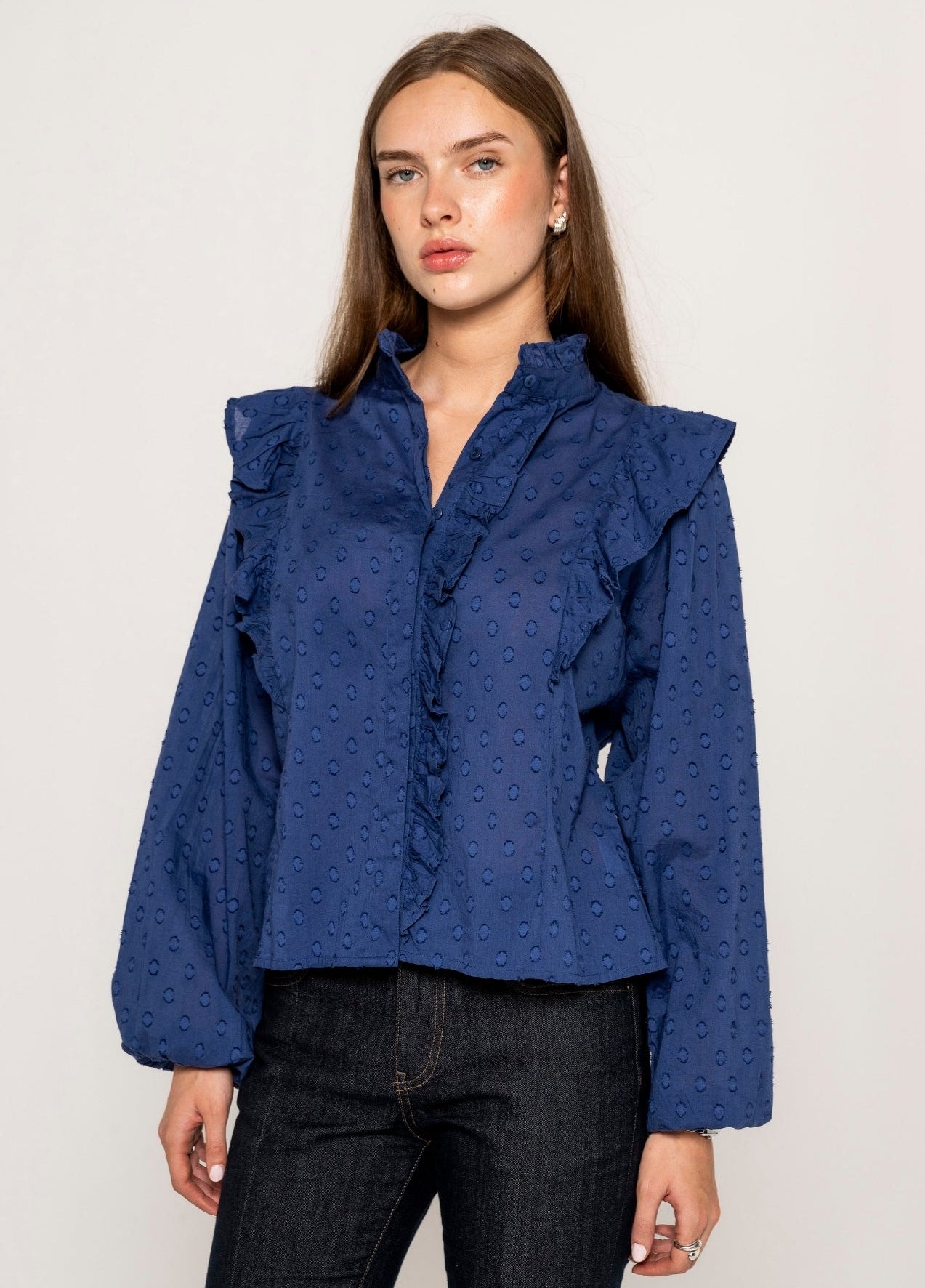 A woman modeling a blue long-sleeve cotton blouse with ruffled details and a textured pattern, paired with dark jeans, against a plain light background.