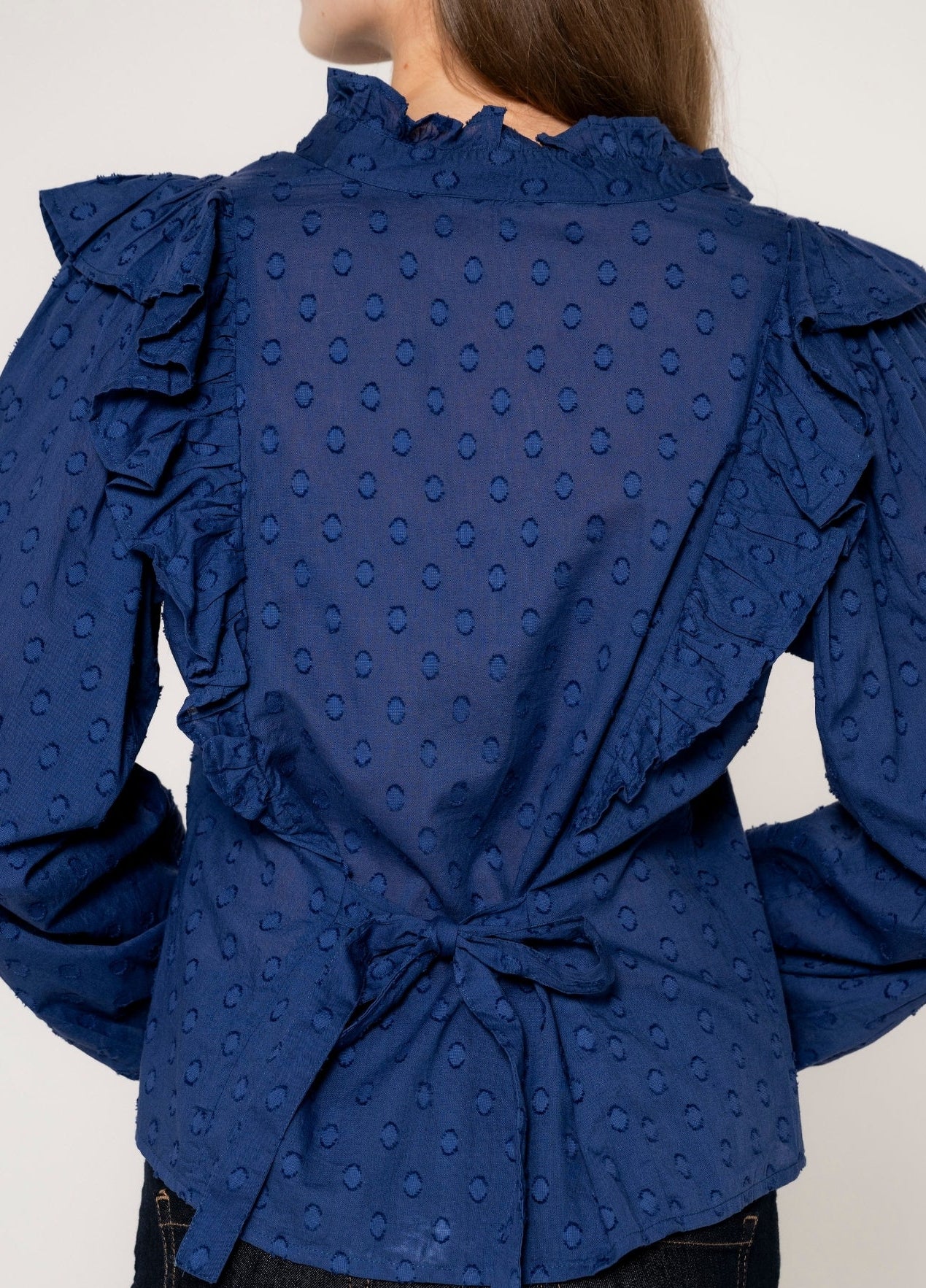 A close-up back view of a woman wearing a royal blue long-sleeve blouse with a textured eyelet pattern, ruffled collar, and ruffled sleeves, tied at the waist.