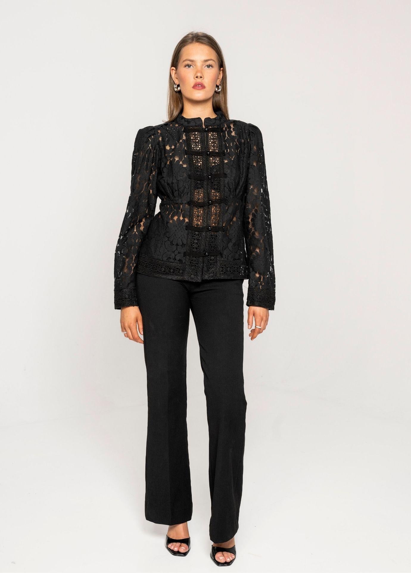Amora Blouse Noir Net: a woman wears a fitted black lace blouse with a stand collar and frog closures down a crochet lace panel. The blouse is styled with black flared trousers and black strappy heels against a plain light grey studio background.