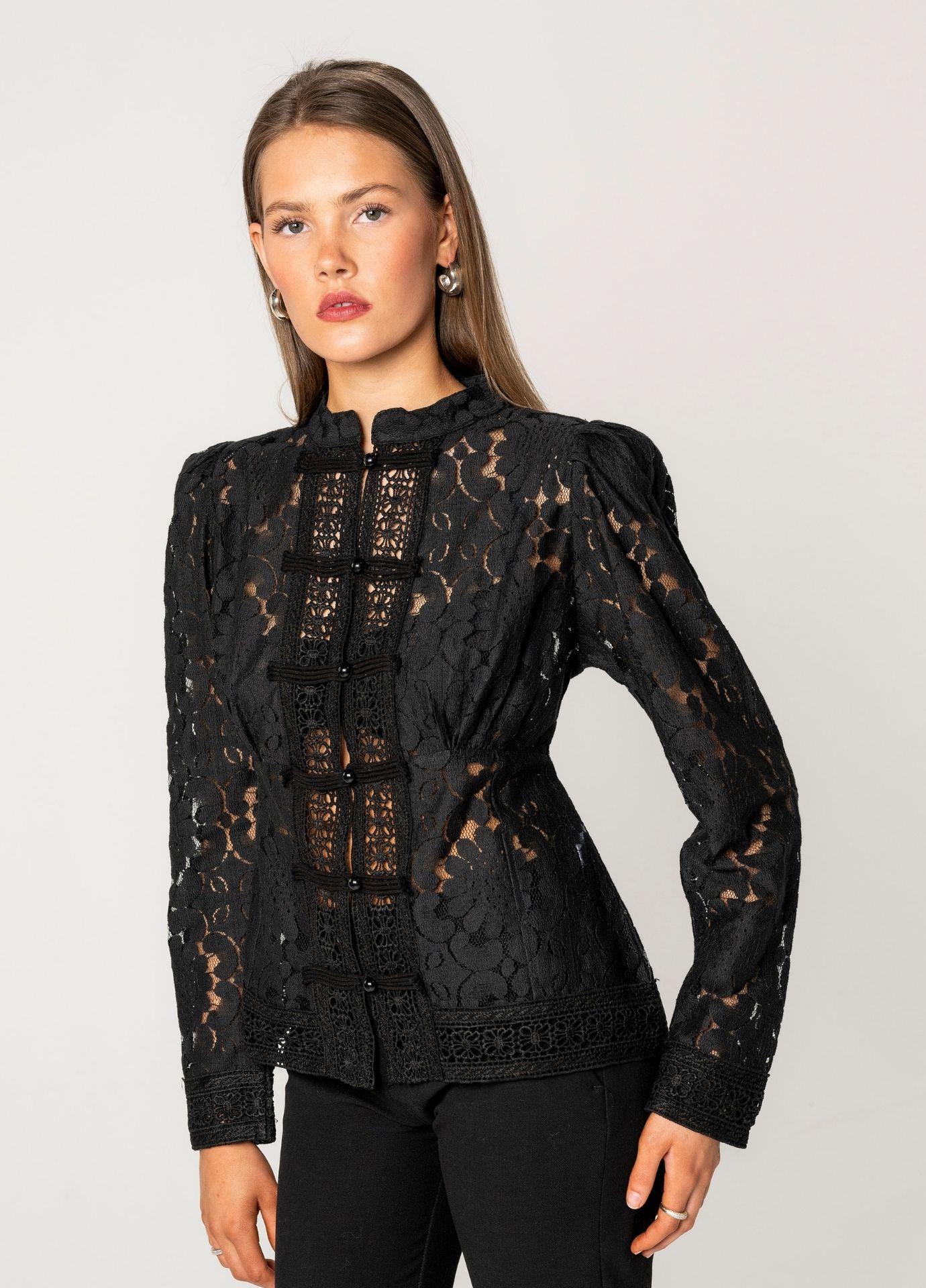 Amora Blouse Noir Net seen from a slight angle, showing the softly puffed shoulder and the gathered seams at the waist. The sheer floral lace lets a hint of skin show through, and the model wears silver hoop earrings against a light grey background.
