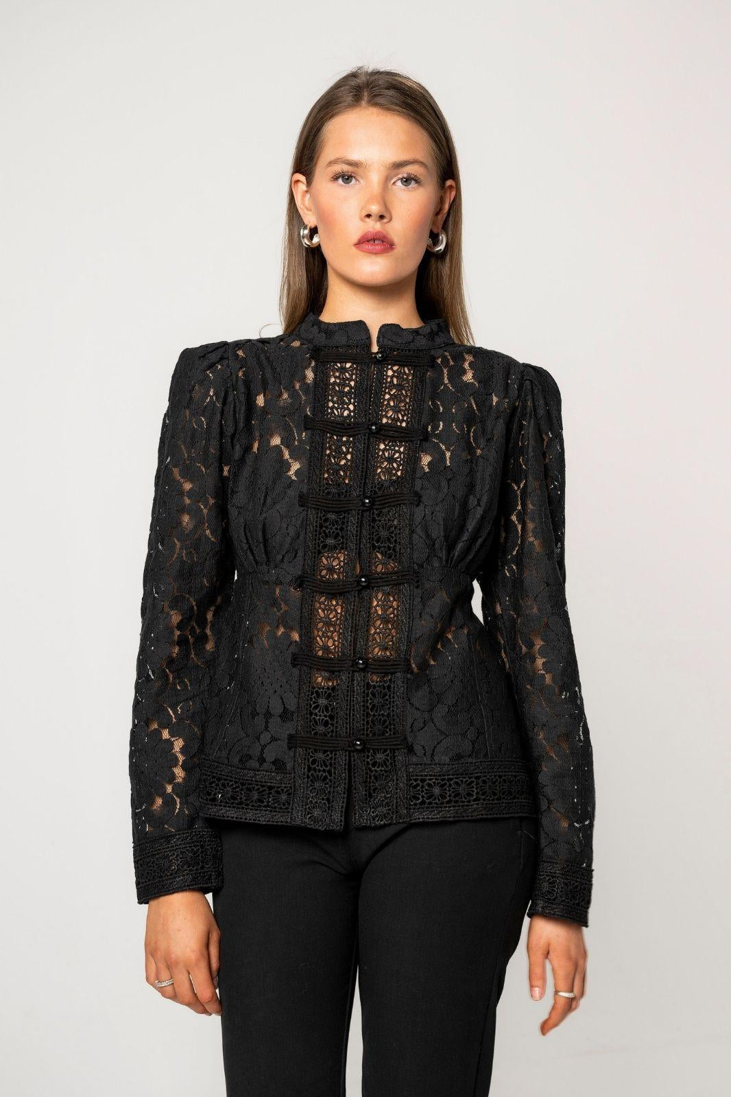 Amora Blouse Noir Net in a close front view. Black frog closures fasten over a vertical panel of crochet lace, and a lace border finishes the hem and cuffs. The model wears black trousers against a plain light grey wall.