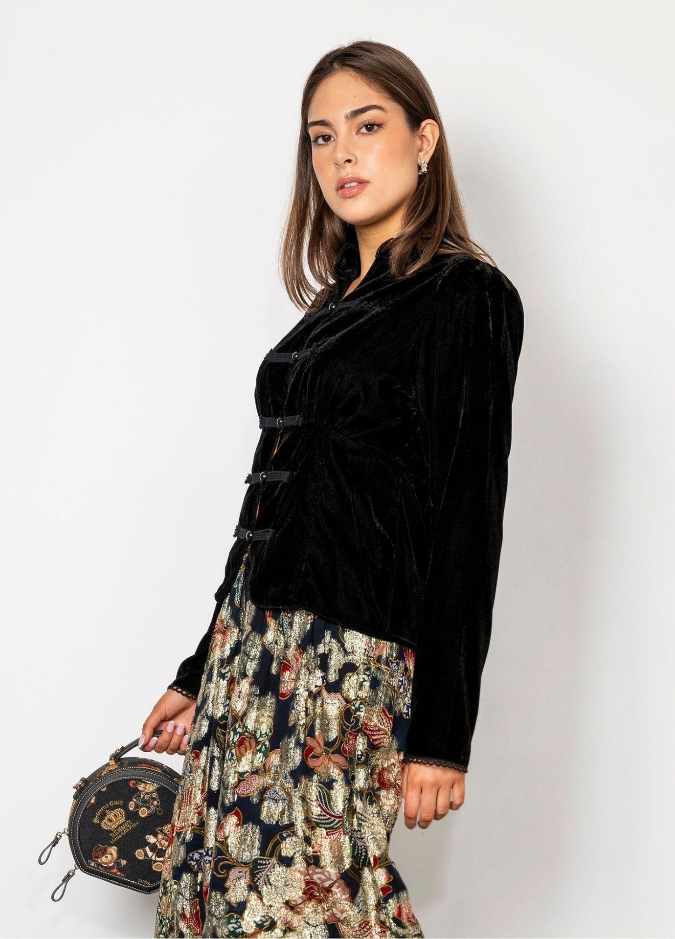 Amora Blouse Velvet Black seen at an angle, showing the frog closures and the puffed shoulders, the model holding a small embroidered round handbag against a white wall.