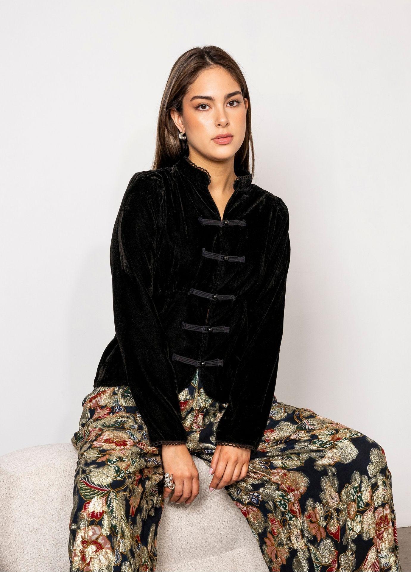 Amora Blouse Velvet Black: a woman sits on a cream chair wearing a fitted black velvet blouse with a stand collar and frog closures, styled with black and gold floral wide leg pants.