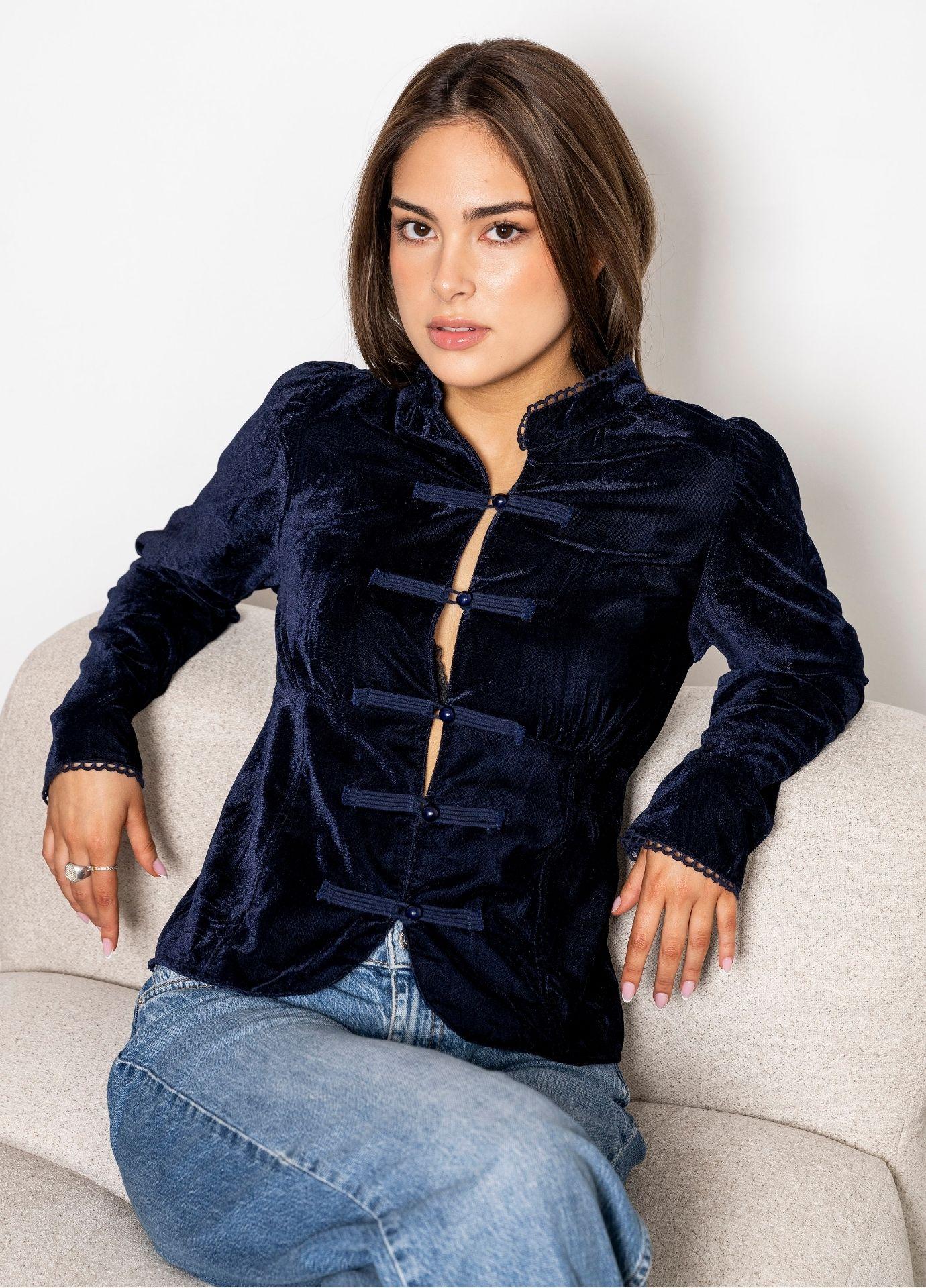 Amora Blouse Velvet Blue Sapphire: a woman sits on a cream upholstered chair wearing a fitted navy velvet blouse with a stand collar and frog closures, paired with light blue jeans against a white studio wall.