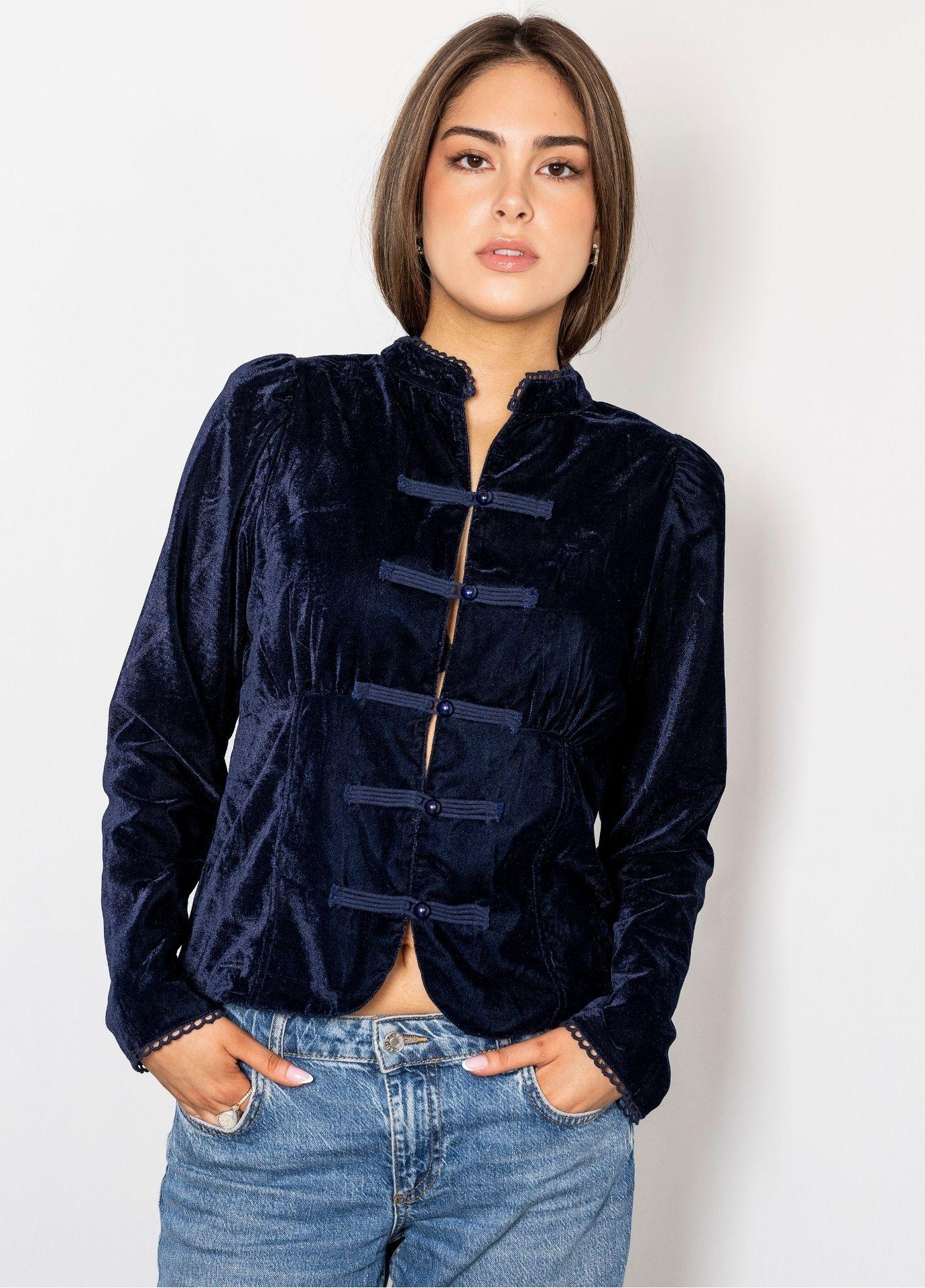 Amora Blouse Velvet Blue Sapphire in a front view. The navy velvet blouse fastens with frog closures and covered buttons, finished with fine lace trim at the collar, and is styled with light blue jeans against a plain white background.
