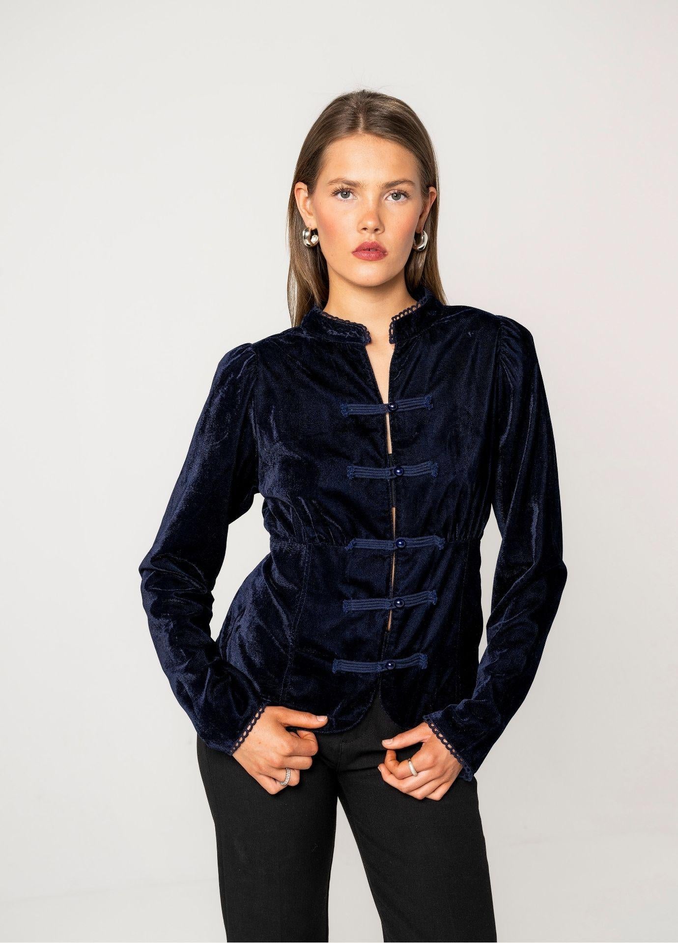 Amora Blouse Velvet Blue Sapphire styled with black trousers. The model stands with her hands in her pockets, showing the puffed shoulders and fitted long sleeves against a light grey studio background.