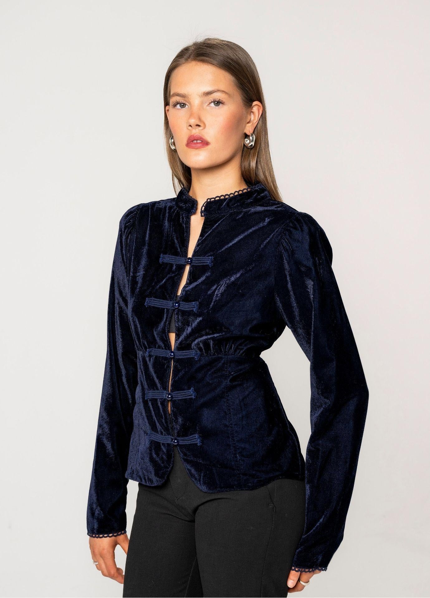 Amora Blouse Velvet Blue Sapphire seen at an angle, showing the shaped waist and the soft sheen of the navy velvet. Styled with black trousers against a light grey background.