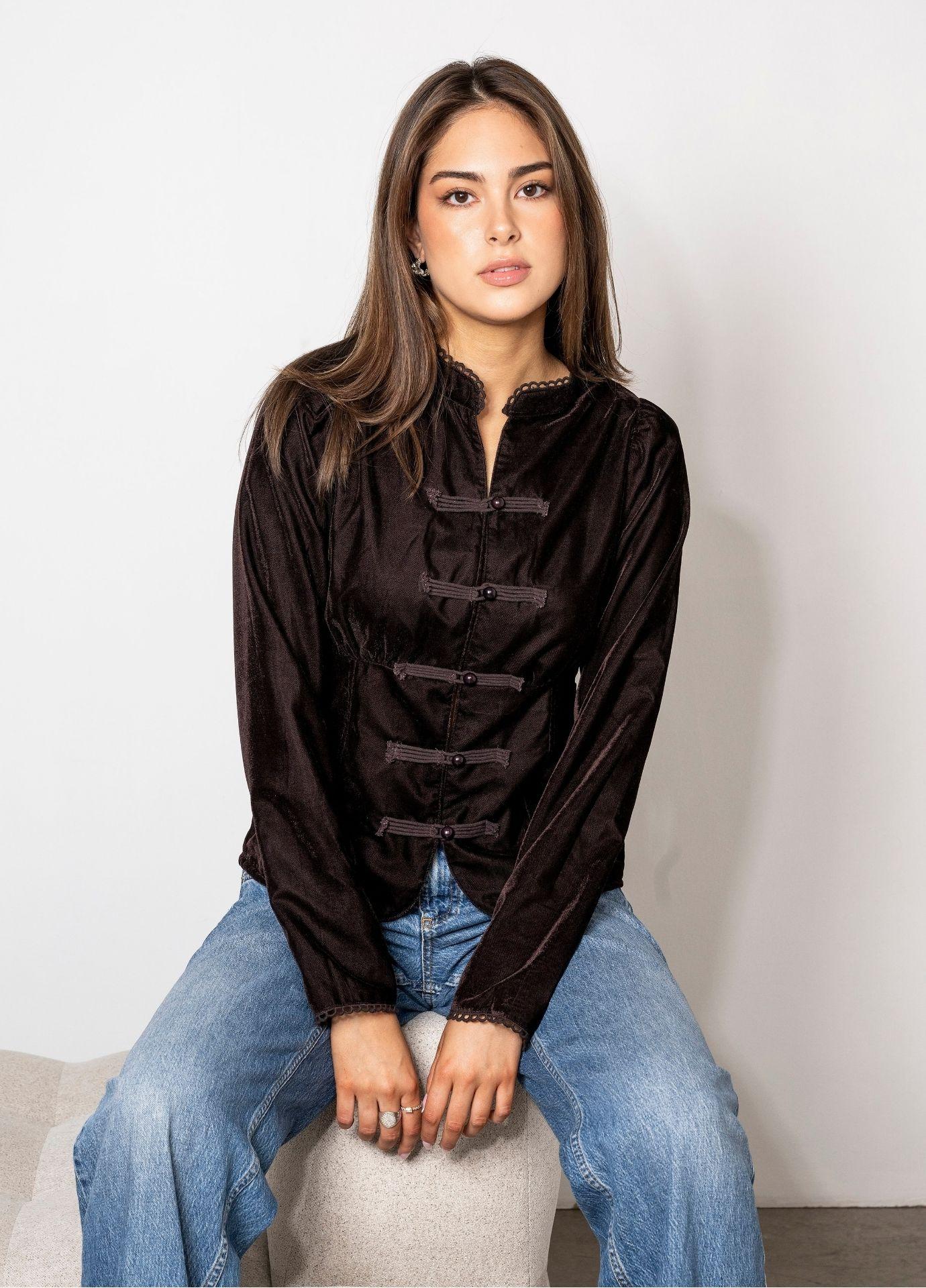 Amora Blouse Velvet Toffee: a woman sits on a cream upholstered chair wearing a fitted toffee brown velvet blouse with a stand collar and frog closures, paired with light blue wide leg jeans against a white wall.