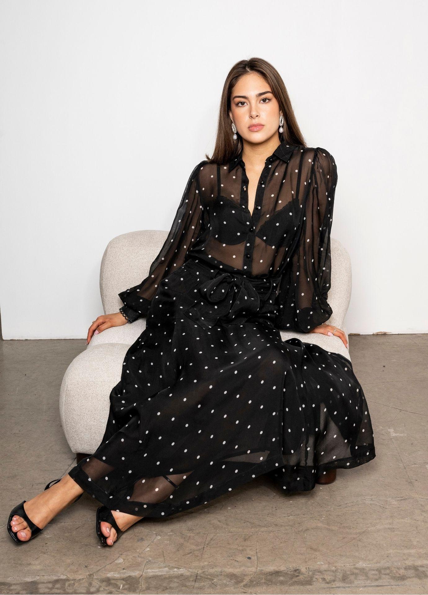 Anaya Blouse Black Pearl worn with the matching Vanja Skirt Black Pearl, a sheer dotted midi skirt tied with a bow at the waist. The model sits on a cream upholstered chair on a concrete floor, wearing black strappy heels.