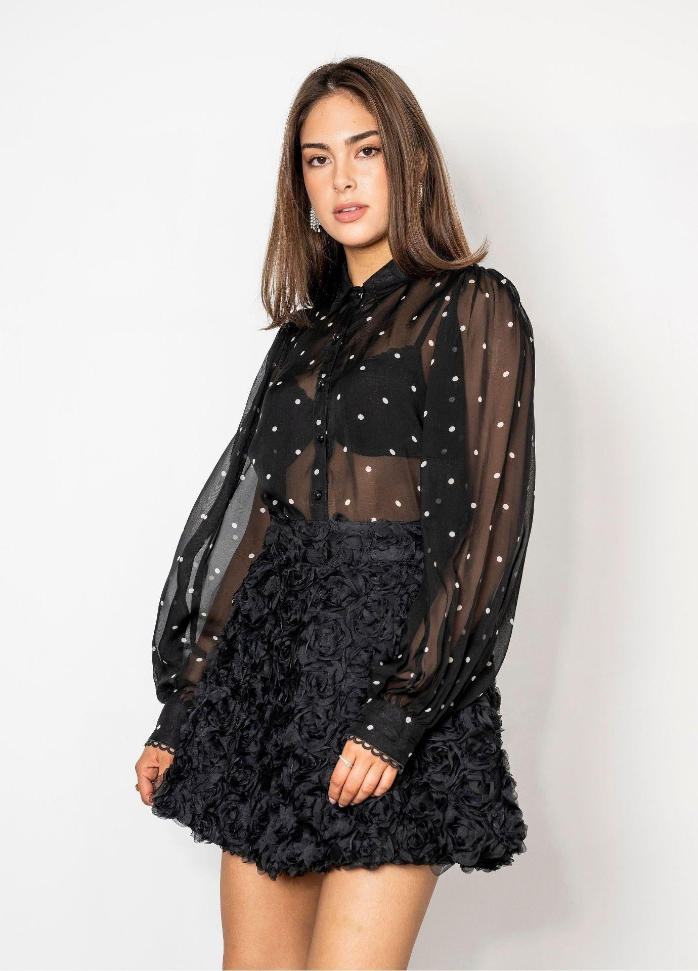 Anaya Blouse Black Pearl styled with a black mini skirt covered in three dimensional rosettes. The sheer dotted blouse has a classic collar and a button front, shown against a plain white studio wall.