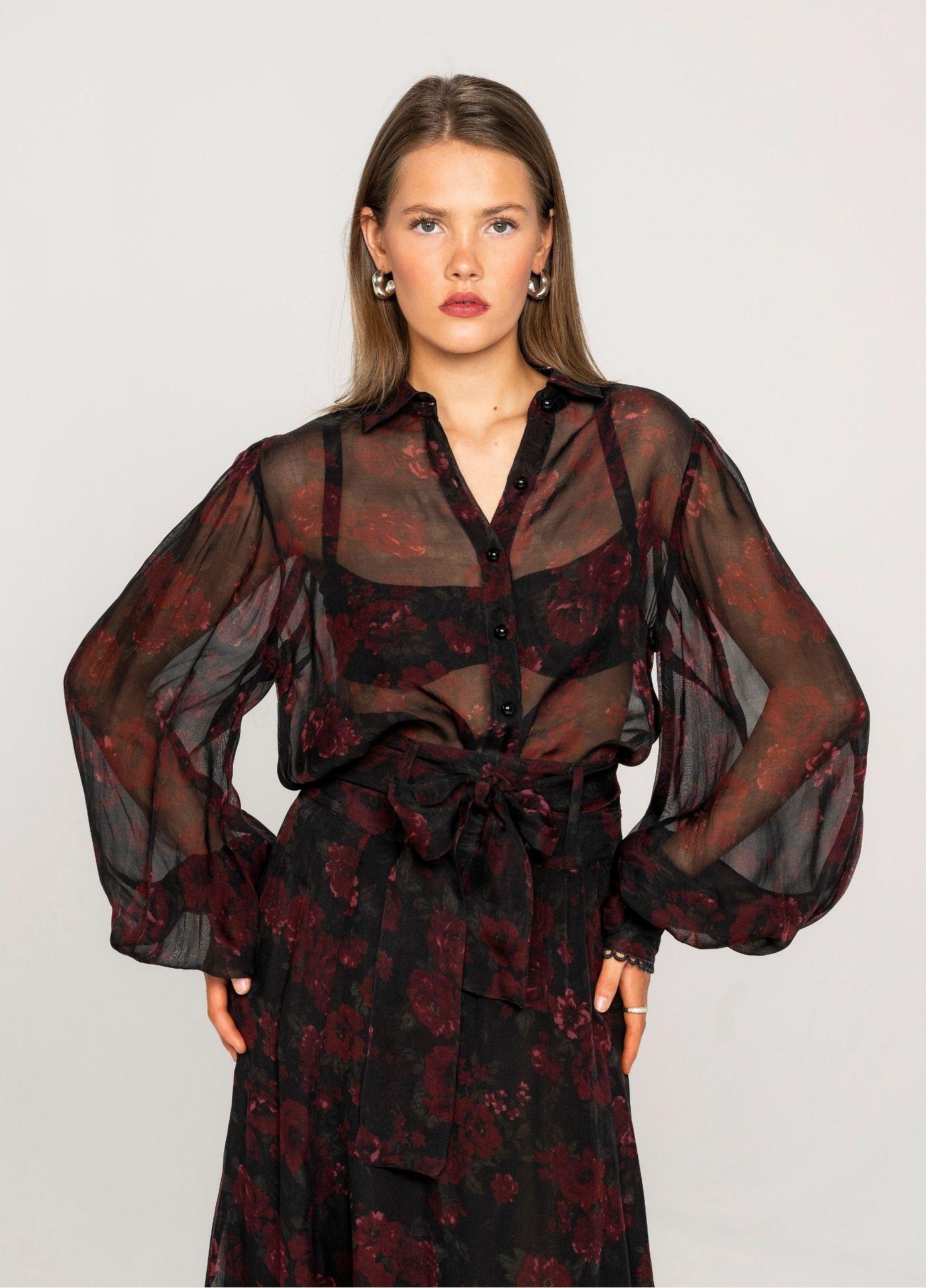 Anaya Blouse Black Romance: a woman wears a sheer black button-down blouse with a deep red floral print and long balloon sleeves, tucked into the matching Vanja Skirt Black Romance. A black bralette shows through, against a plain light grey studio background.