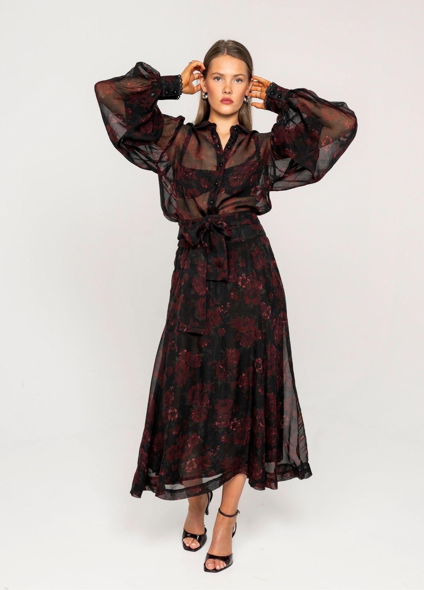 Anaya Blouse Black Romance in a full length view with the model's hands raised to her hair, showing the volume of the sheer balloon sleeves. Styled with the matching floral midi skirt and black strappy heels on a light grey background.