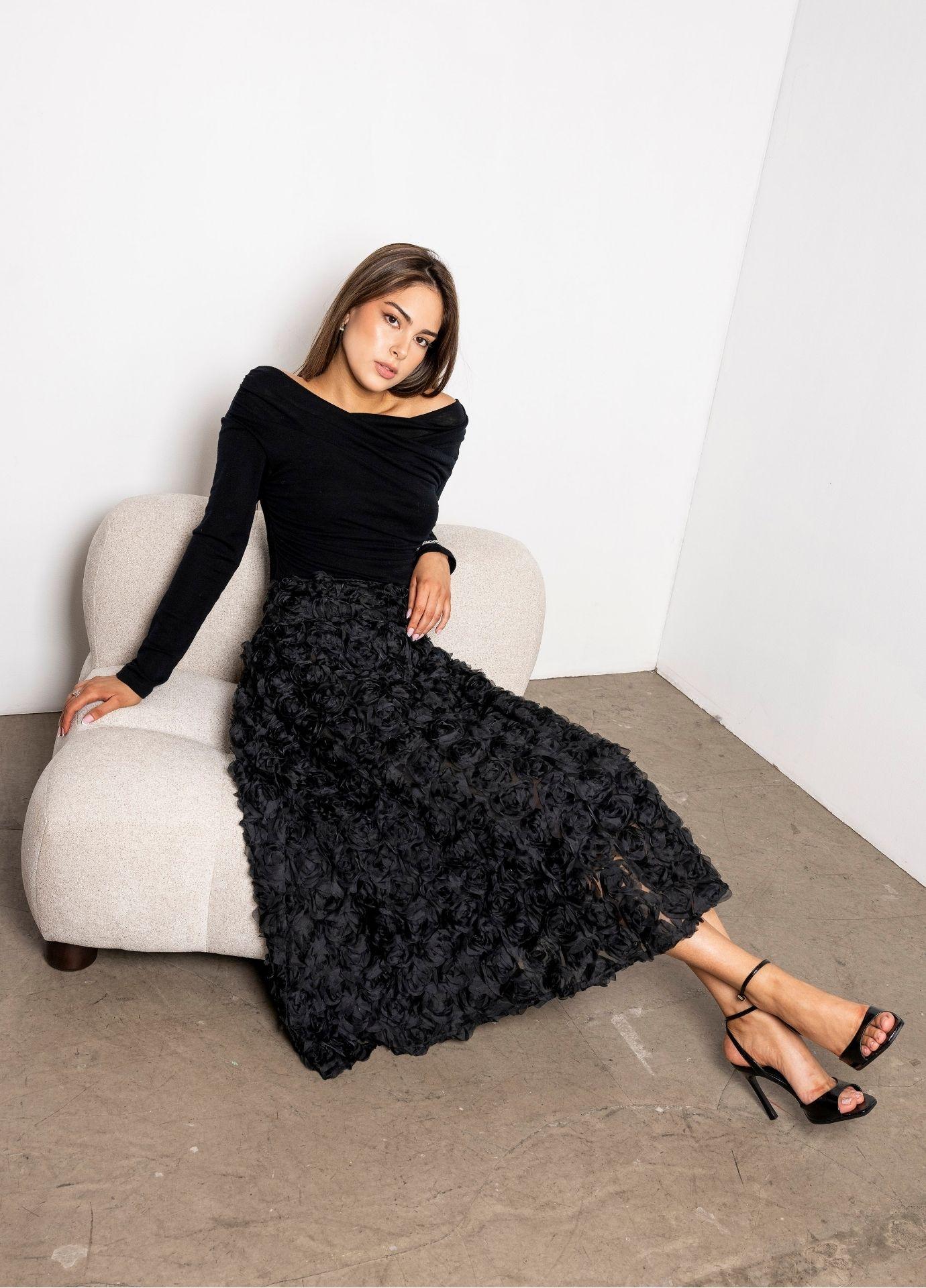 Astrid Skirt Rosette: a woman sits on a cream upholstered chair wearing a full black midi skirt covered in three dimensional rosettes, styled with a black long sleeve top and black heels on a concrete floor.