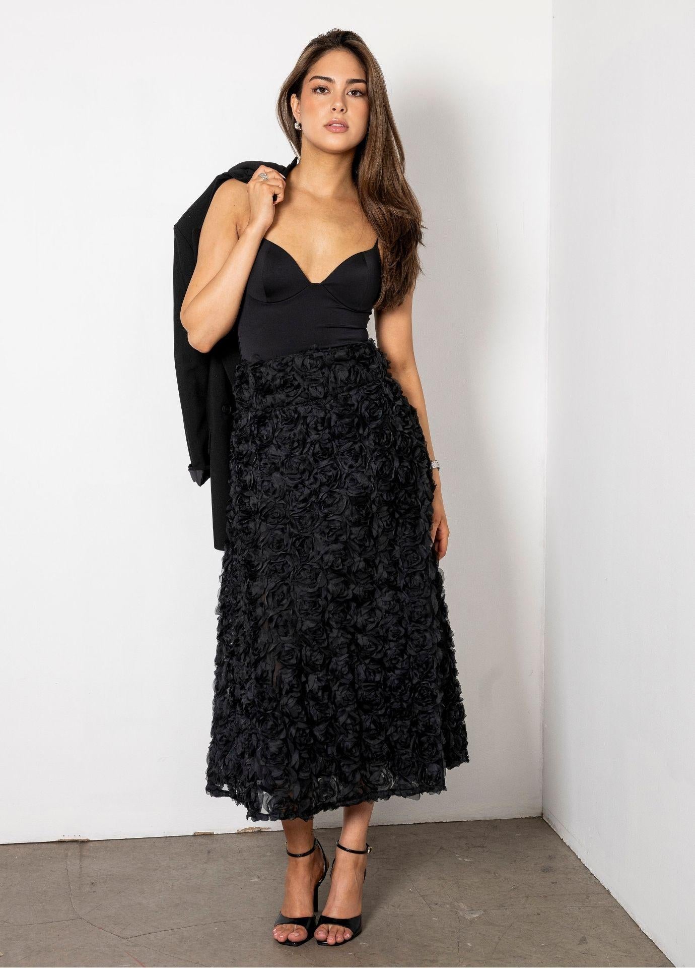 Astrid Skirt Rosette in a full length front view, styled with a black bustier top and a black blazer over the shoulder. The textured rosette skirt falls to mid calf against a white wall.