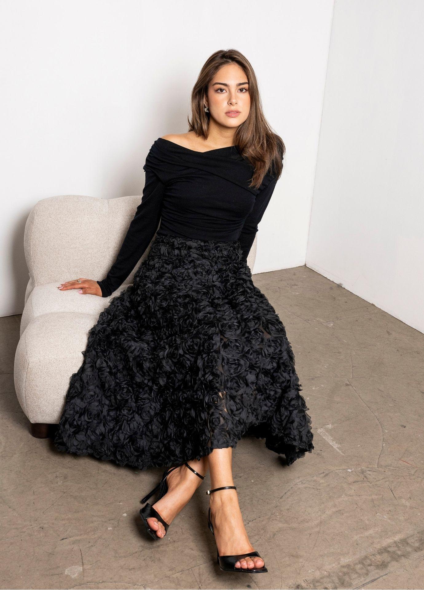 Astrid Skirt Rosette in a front view, the model seated on a cream chair in a black top and strappy heels against a white studio wall.