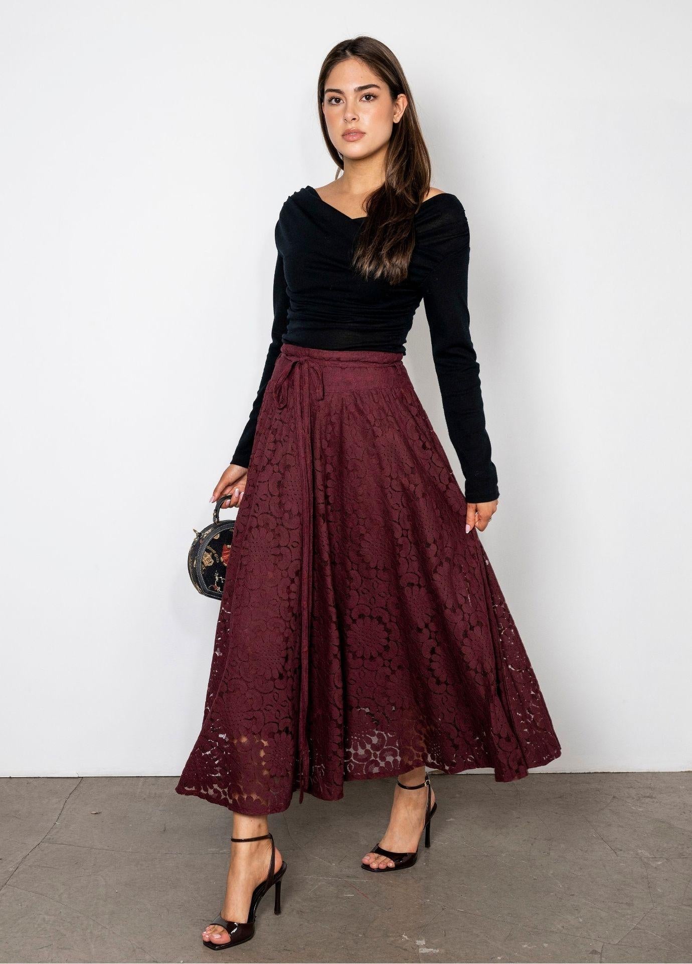 Astrid Skirt Vintage Wine: a woman wears a full wine red floral lace midi skirt with a matching tie belt, styled with a black long sleeve top and black heels against a white wall.