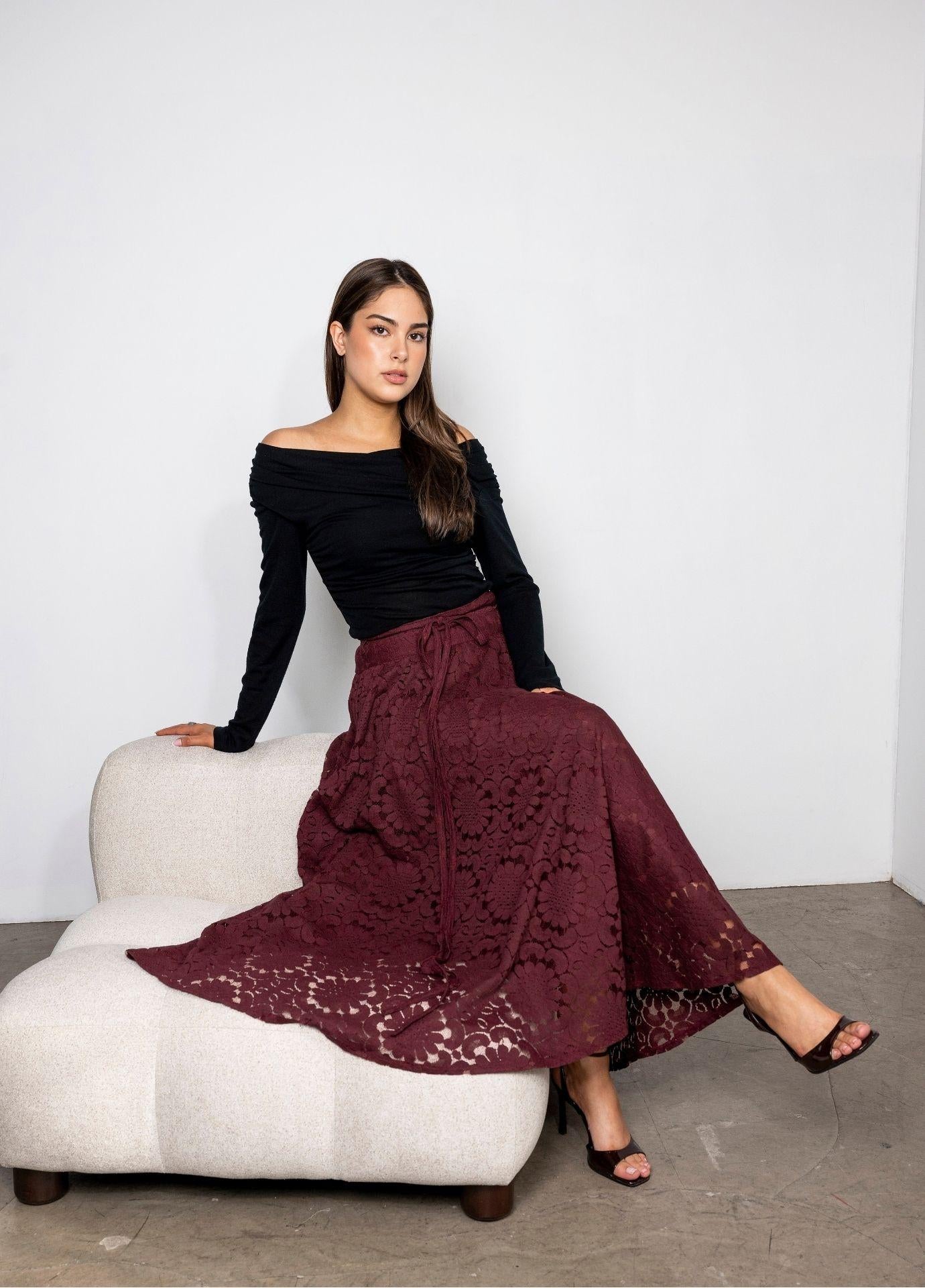 Astrid Skirt Vintage Wine with the model seated on a cream upholstered chair and the lace skirt spread around her, styled with a black off shoulder top.