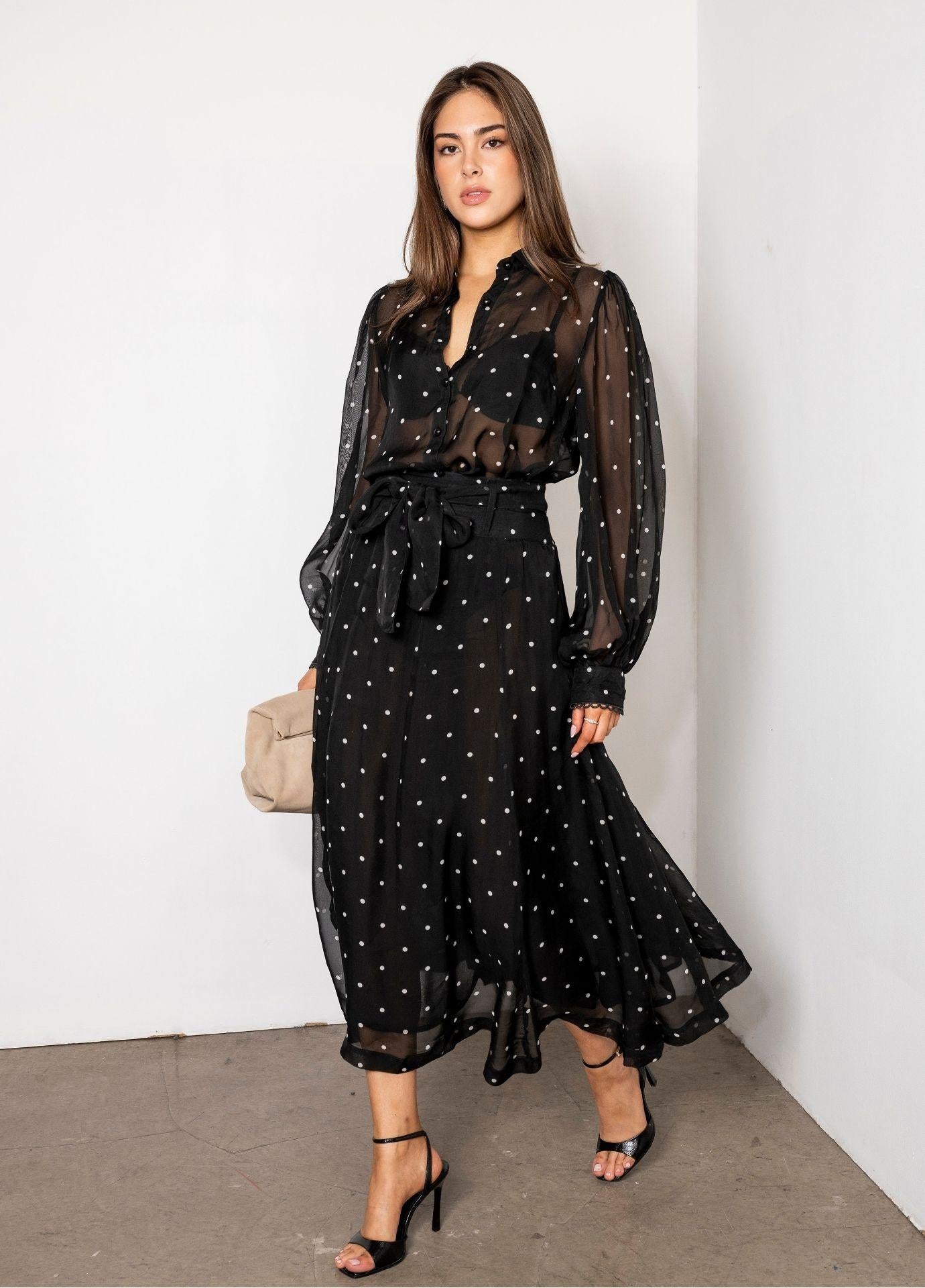 Vanja Skirt Black Pearl in motion, the sheer dotted skirt swinging below the knee over a shorter lining. Styled with the matching Anaya Blouse Black Pearl, black heels and a beige handbag against a white wall.