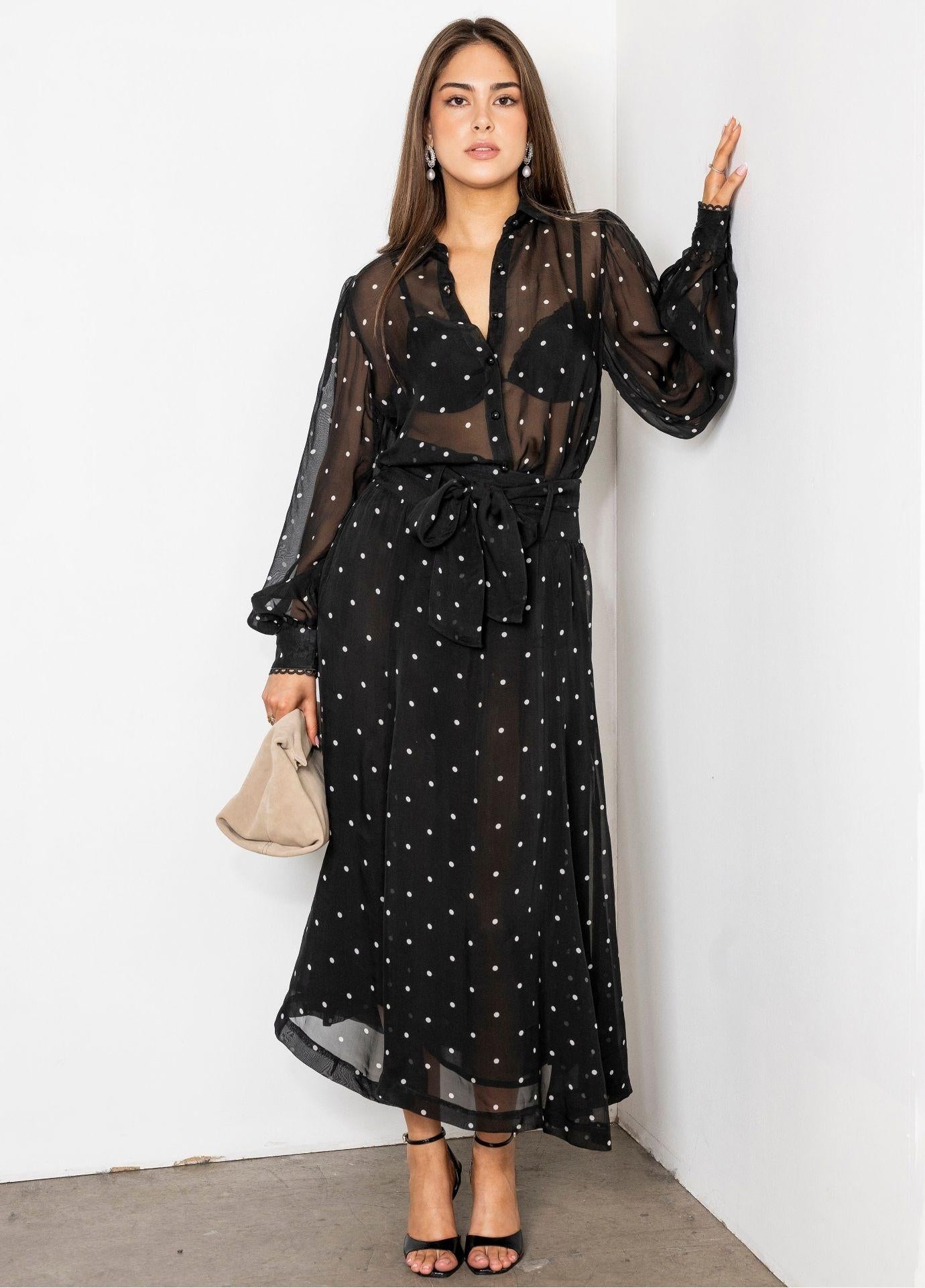 Vanja Skirt Black Pearl in a full length view, worn with the matching sheer dotted blouse and a black bralette. The model leans against a white wall holding a beige handbag, showing the flowy midi length and the tie belt.