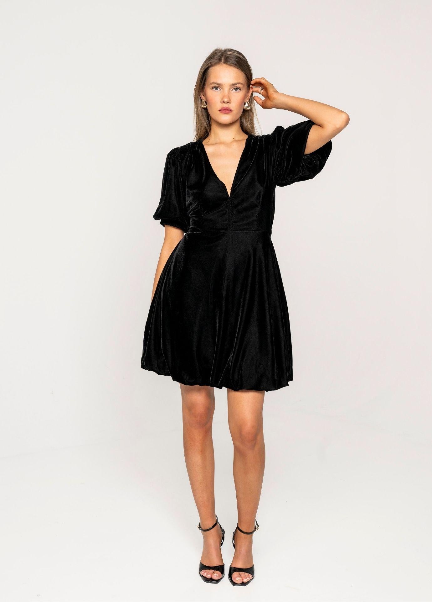Blake Dress Velvet Black in a studio view with one hand raised to her hair, showing the balloon skirt and the short puff sleeves against a light grey background.