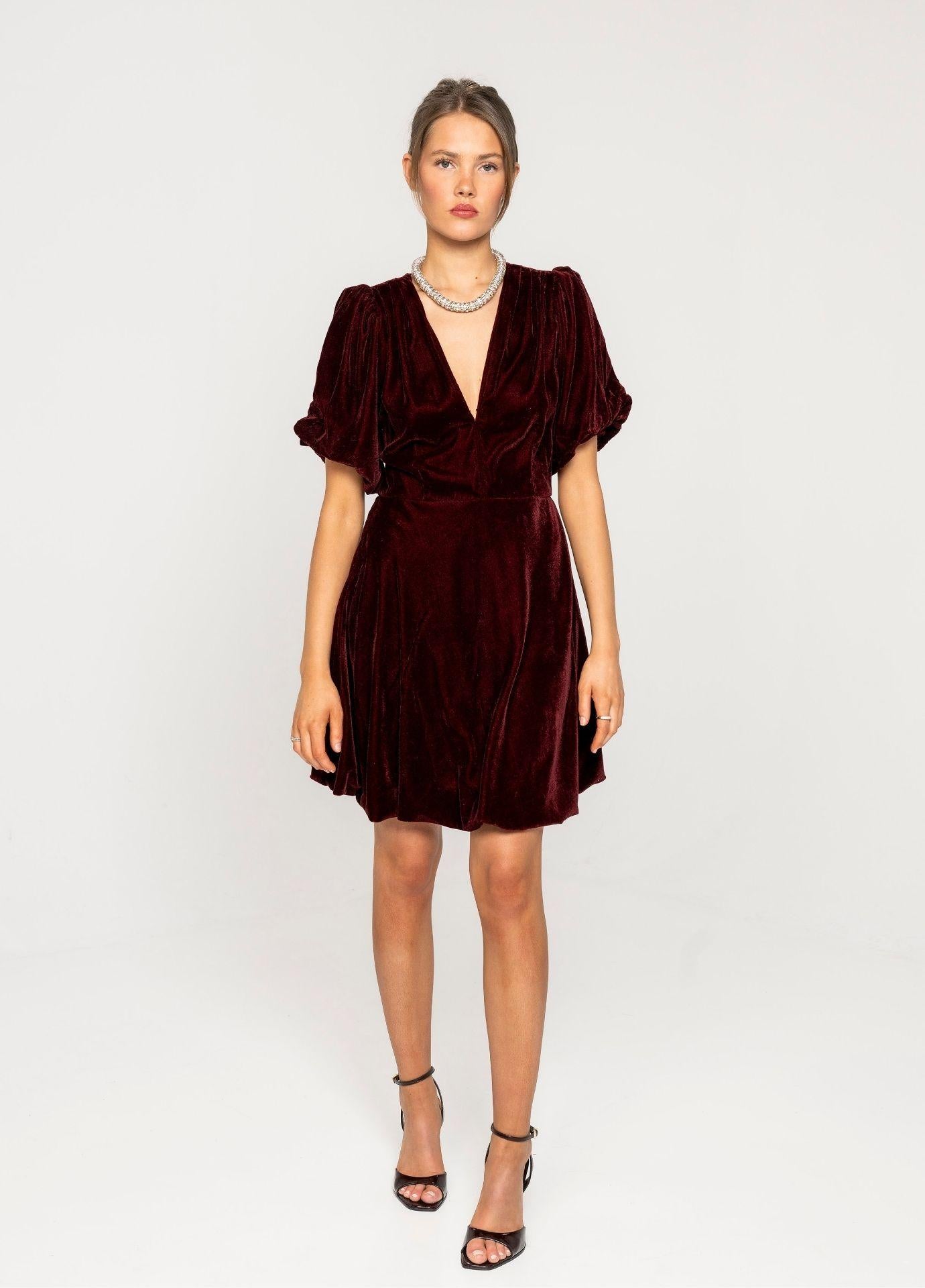 Blake Dress Velvet Bordeaux in a full length front view in a light grey studio. The deep red velvet mini dress with puff sleeves is styled with black strappy heels.