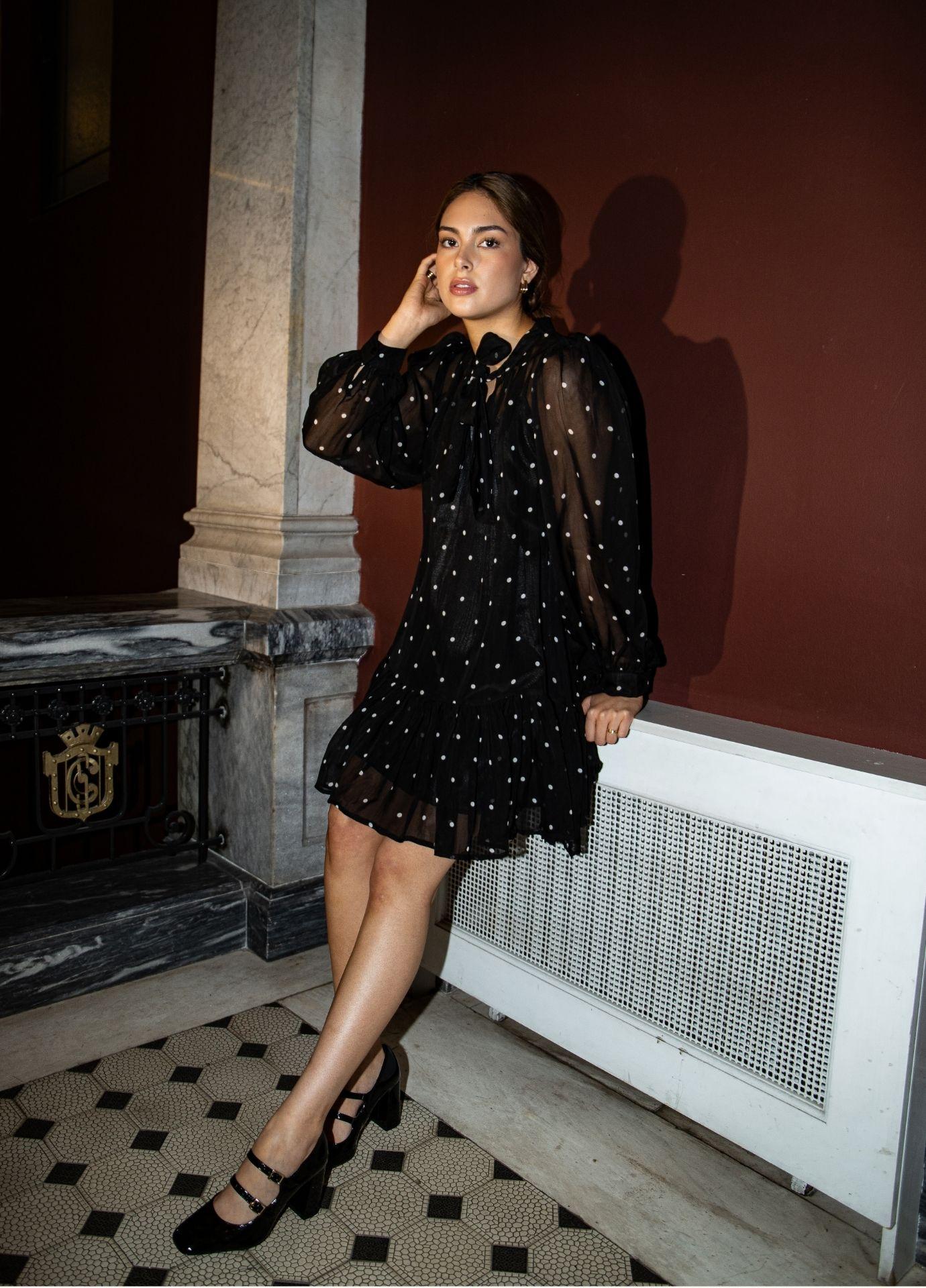 Chloe Dress Black Pearl: a woman sits against a radiator in a marble hallway, wearing a sheer black mini dress with small white dots, a tie at the neck and long sheer balloon sleeves, with black Mary Jane heels.