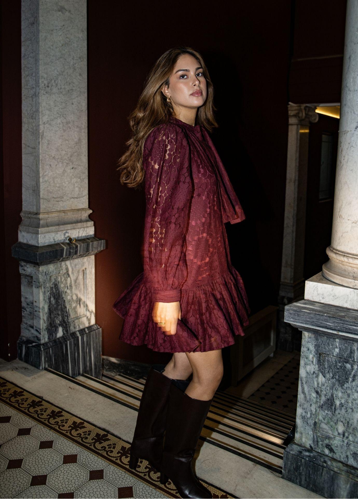 Chloe Dress Vintage Wine: a woman wears a wine red floral lace mini dress with long sleeves, a tie at the neck and a ruffled hem, styled with black knee high boots in a marble hallway.