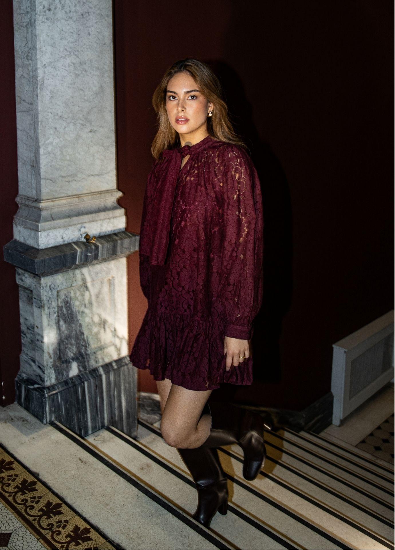 Chloe Dress Vintage Wine in a side view, showing the tie neckline and the sheer floral lace sleeves against dark red walls and marble columns.