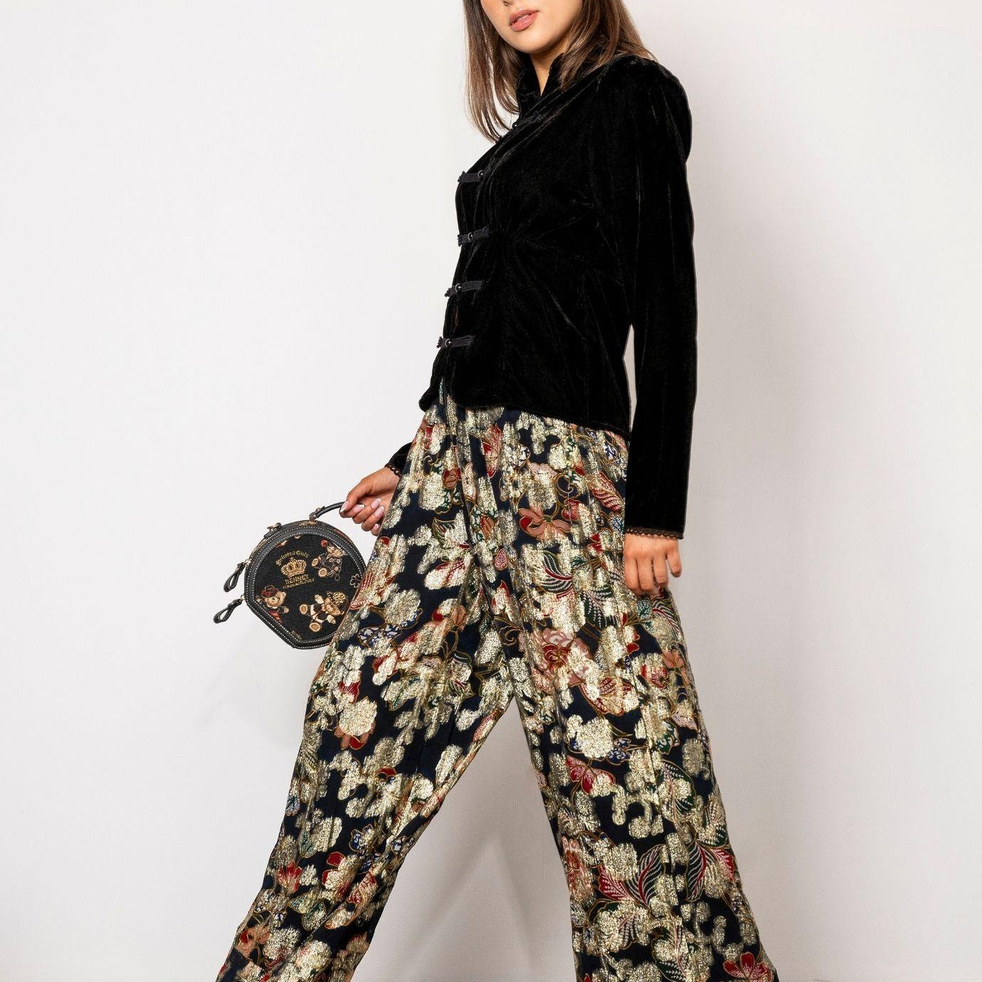 Elida Pant Golden Night: a woman wears high waisted wide leg pants in black with a golden floral pattern, styled with a black velvet blouse with frog closures, black heels and a small embroidered round handbag against a white studio wall.