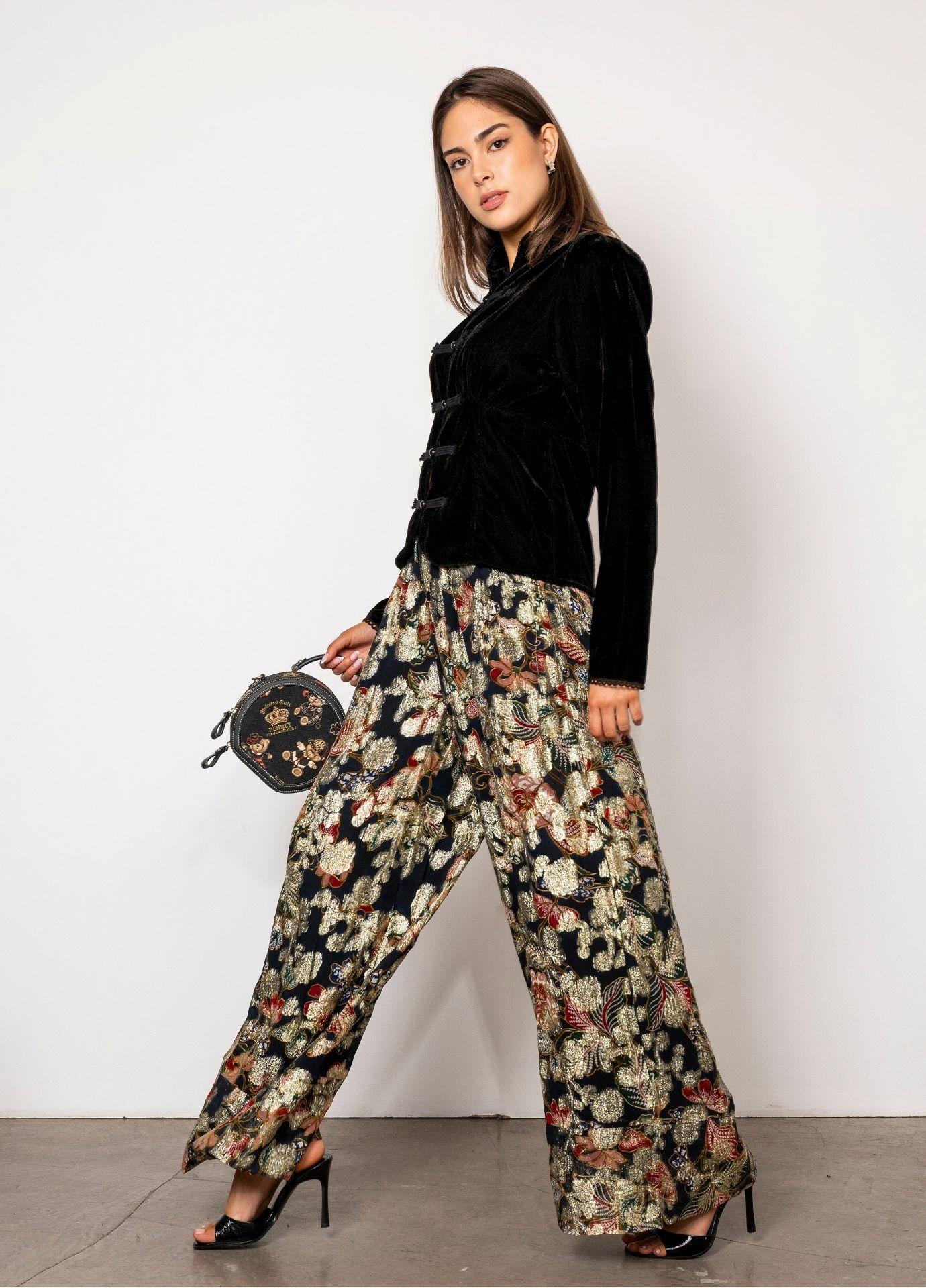 Elida Pant Golden Night: a woman wears high waisted wide leg pants in black with a golden floral pattern, styled with a black velvet blouse with frog closures, black heels and a small embroidered round handbag against a white studio wall.