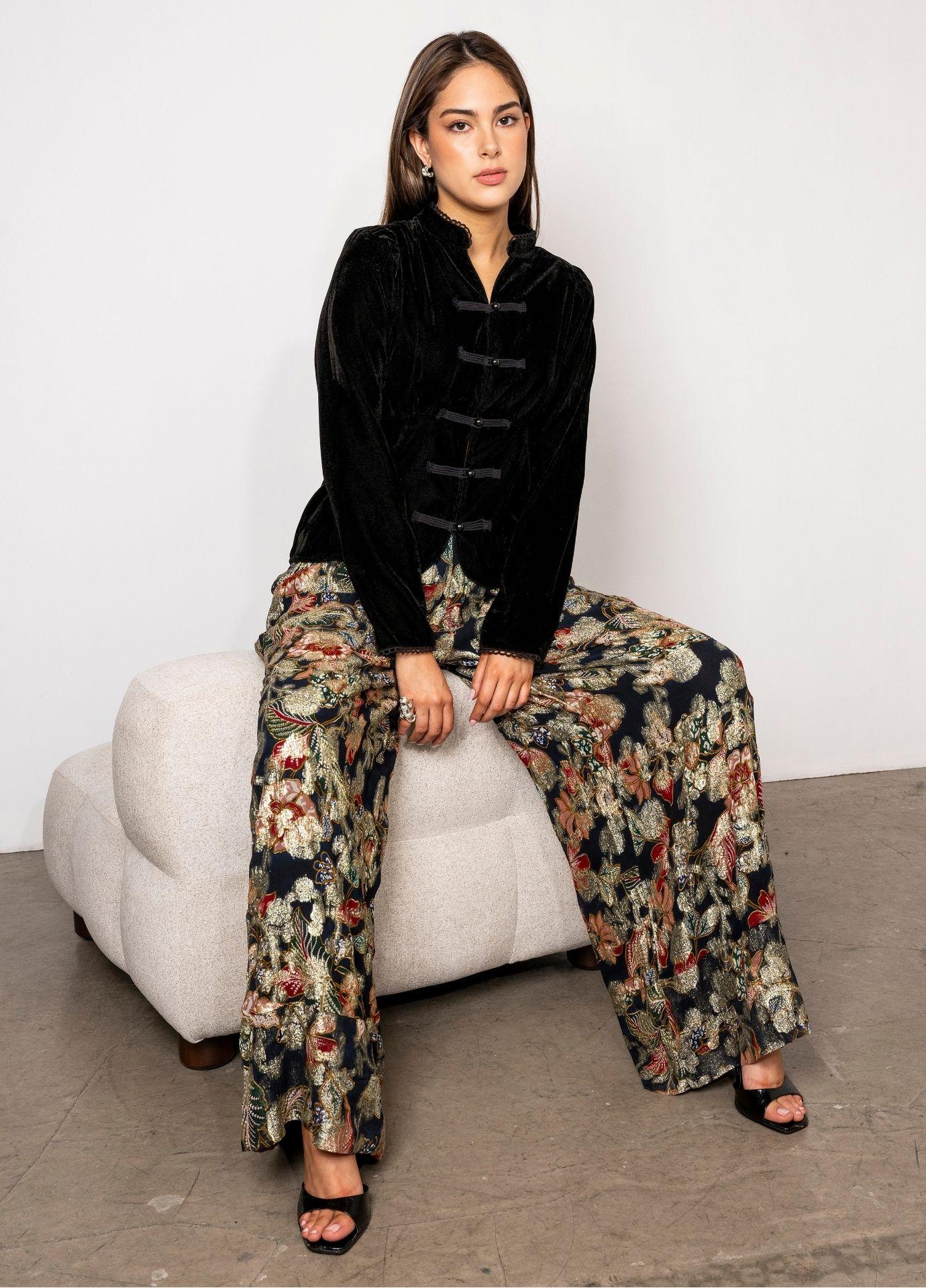 Elida Pant Golden Night with the model seated on a cream upholstered chair, the floral wide leg pants paired with a black velvet blouse.