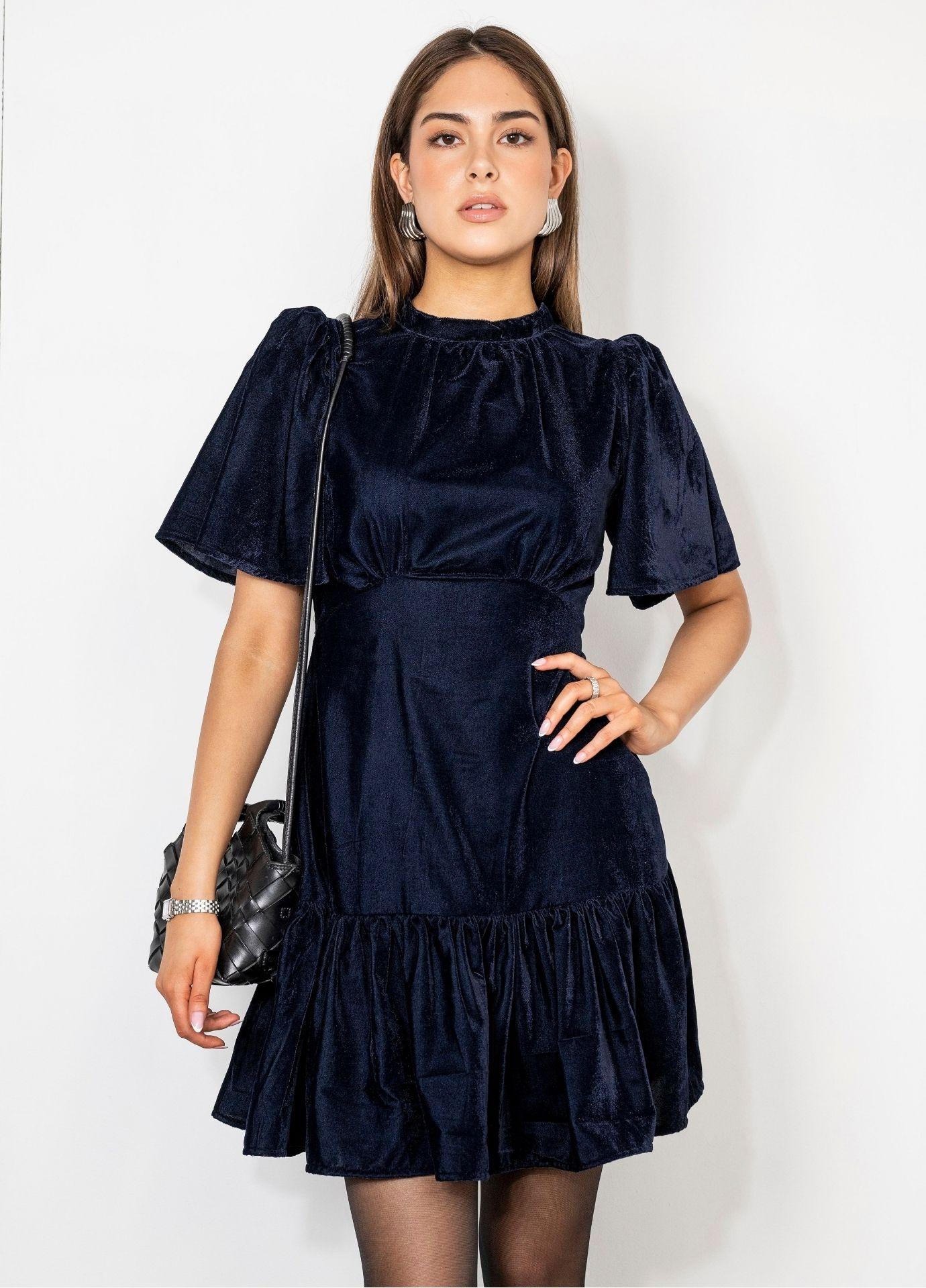 Jolie Dress Velvet Blue Sapphire: a woman wears a navy velvet mini dress with a high gathered neckline, short flared sleeves and a ruffled hem, one hand on her hip and a black quilted bag on her shoulder against a white wall.