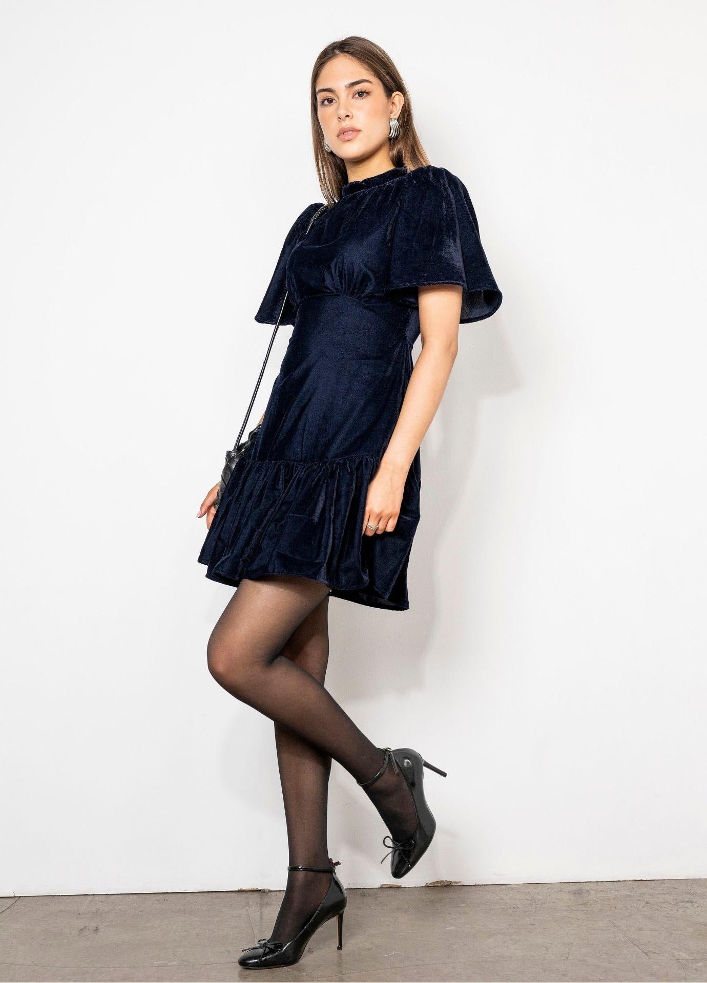 Jolie Dress Velvet Blue Sapphire in a full length side view, styled with sheer black tights and black shoes, showing the ruffled mini hem.