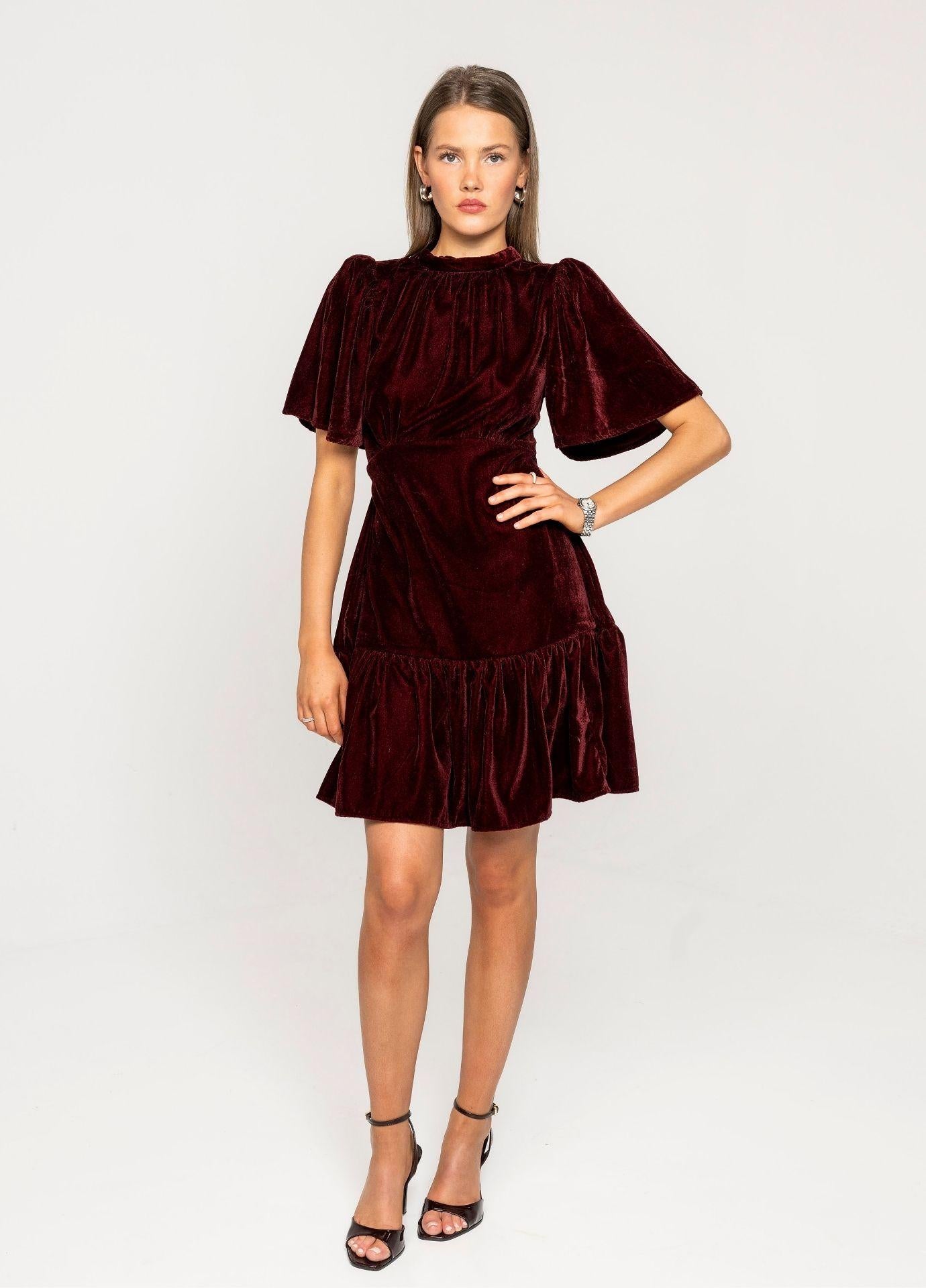 Jolie Dress Velvet Bordeaux in a full length front view in a light grey studio, one hand on the hip, styled with black strappy heels.