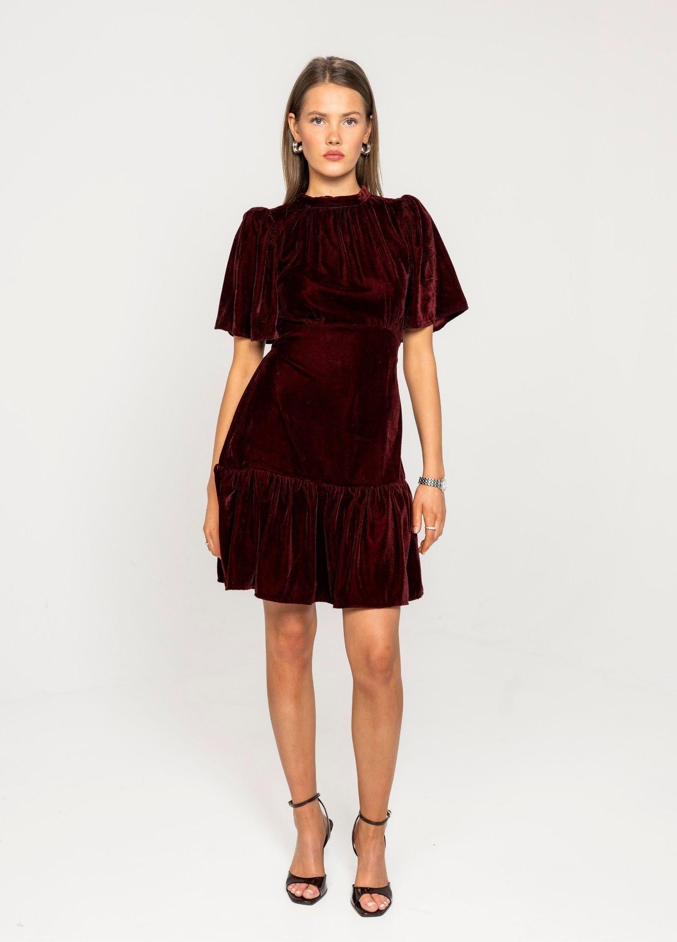 Jolie Dress Velvet Bordeaux in a studio front view, showing the fitted bodice, the short flared sleeves and the ruffled hem of the red velvet mini dress.