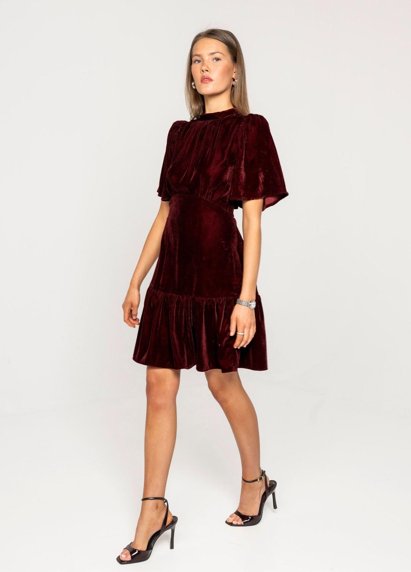 Jolie Dress Velvet Bordeaux in a studio view mid step, the ruffled hem moving with black strappy heels against a light grey background.