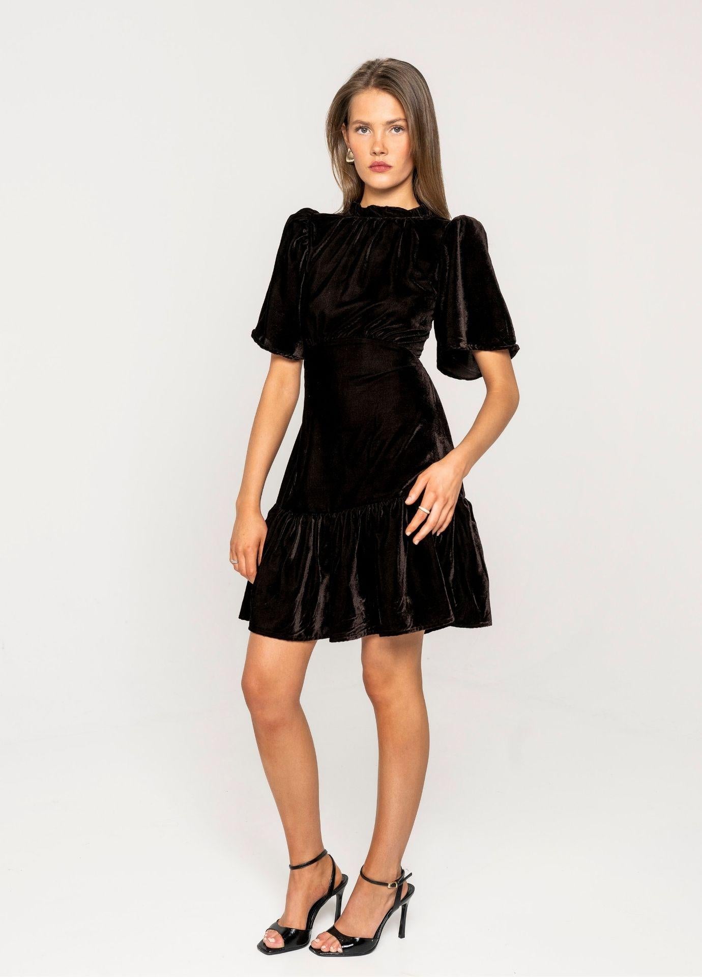 Jolie Dress Velvet Toffee: a woman wears a toffee brown velvet mini dress with a high gathered neckline, short loose sleeves and a ruffled hem, standing in a light grey studio in black strappy heels.