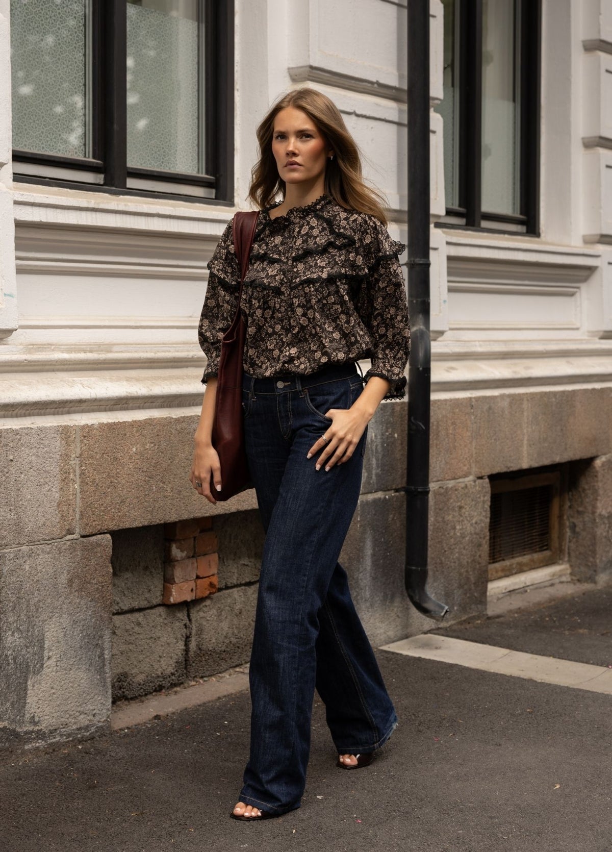 A woman stands on a city sidewalk wearing a dark floral long-sleeve blouse with ruffled detailing, dark blue wide-leg jeans, and carrying a dark brown shoulder bag.