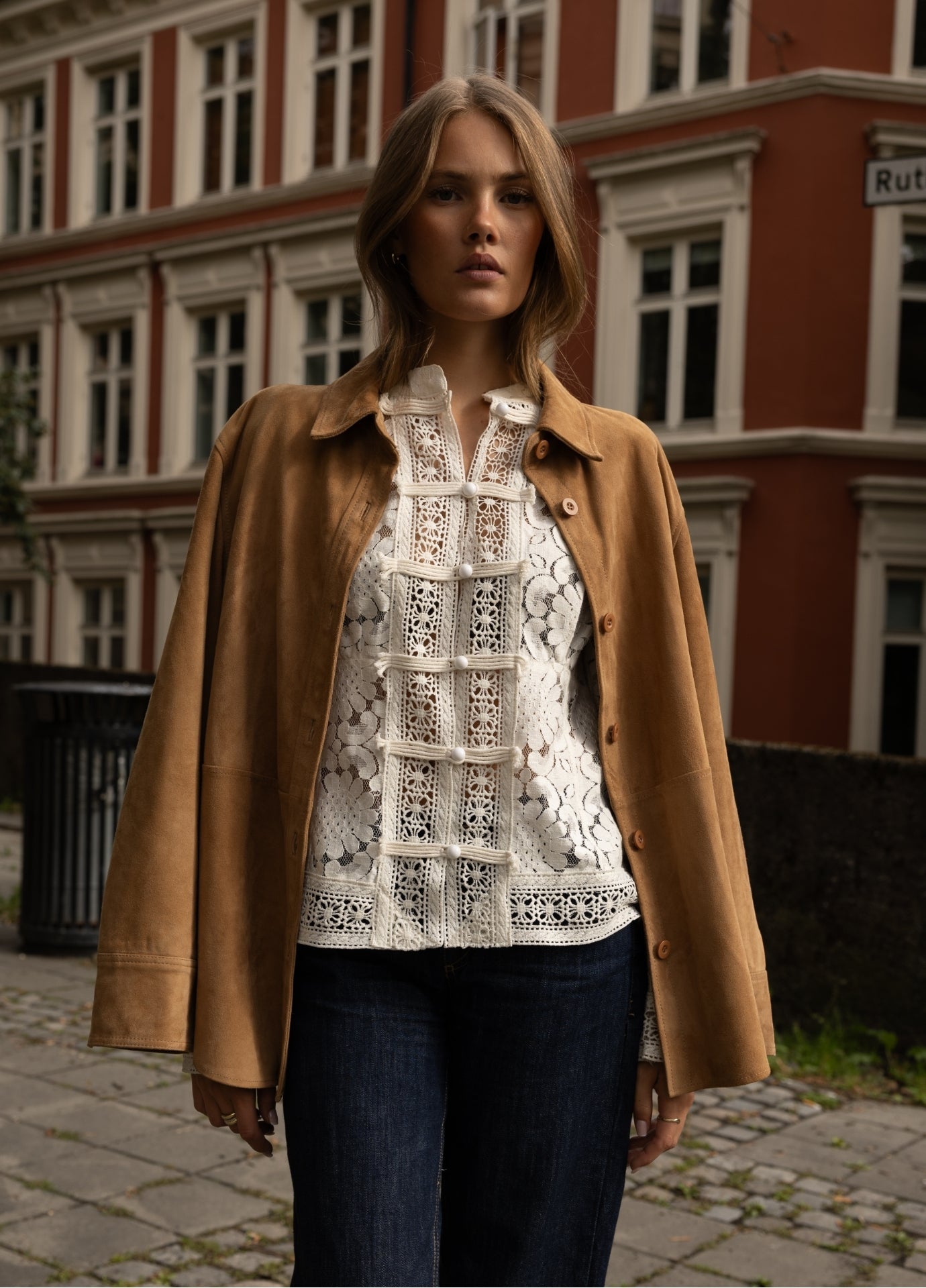 A woman stands outdoors in front of a red brick building, wearing a camel brown coat over a white lace blouse and dark blue jeans.