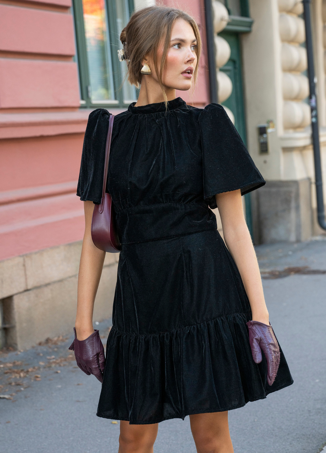 Jolie Dress Velvet Black in a front view on a city street, the fitted velvet bodice and short sleeves paired with burgundy leather gloves.