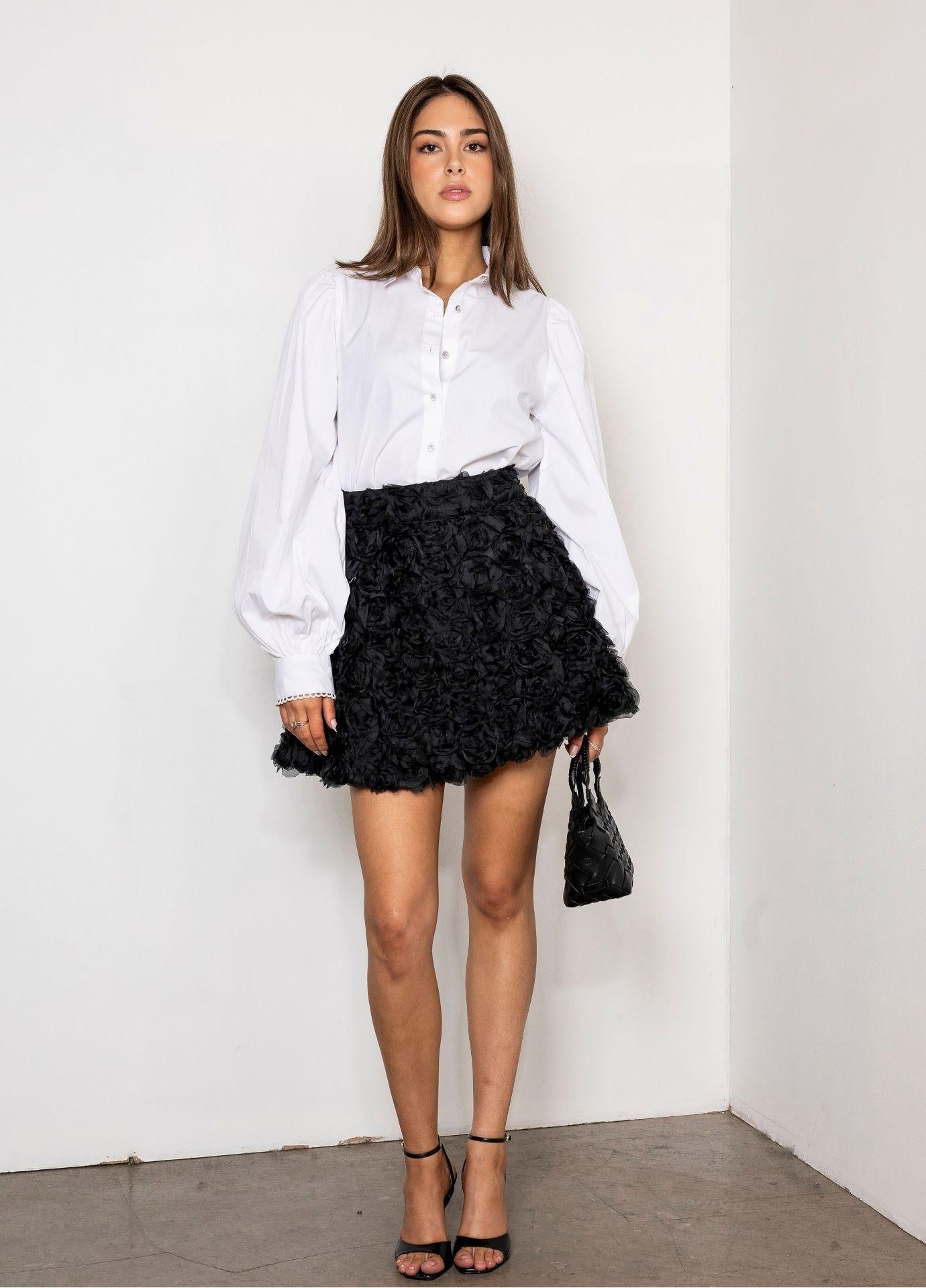 Nina Skirt Rosette: a woman wears a black balloon mini skirt covered in three dimensional rosettes, styled with a crisp white shirt with balloon sleeves and a small black handbag against a white wall.