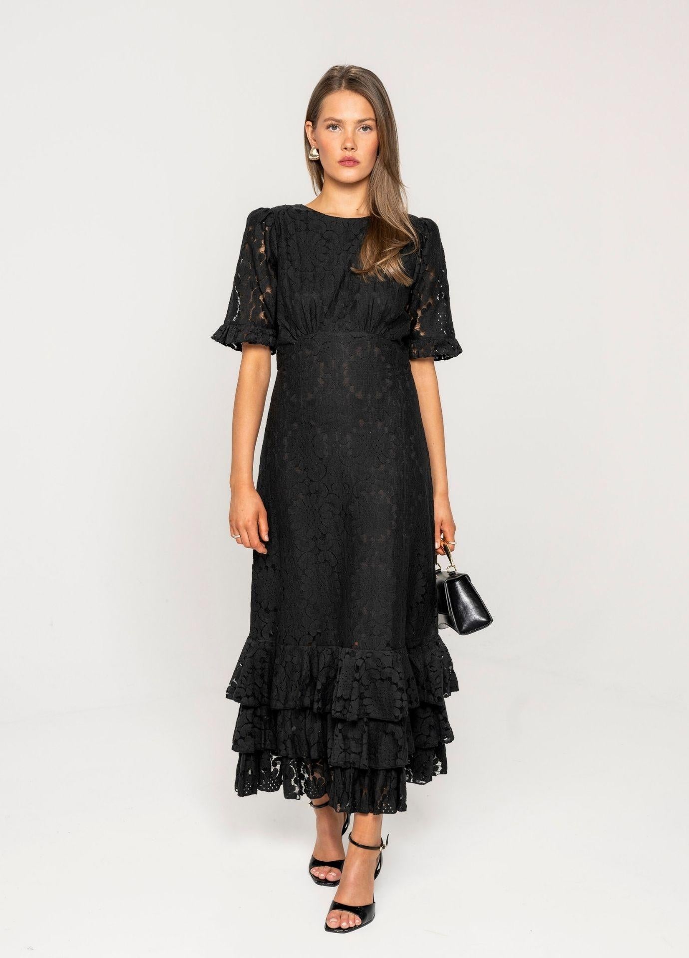 Odette Dress Noir Net: a woman wears a long black lace dress with a round neckline, short ruffled puff sleeves and layered ruffles at the hem, holding a small black handbag in a light grey studio.