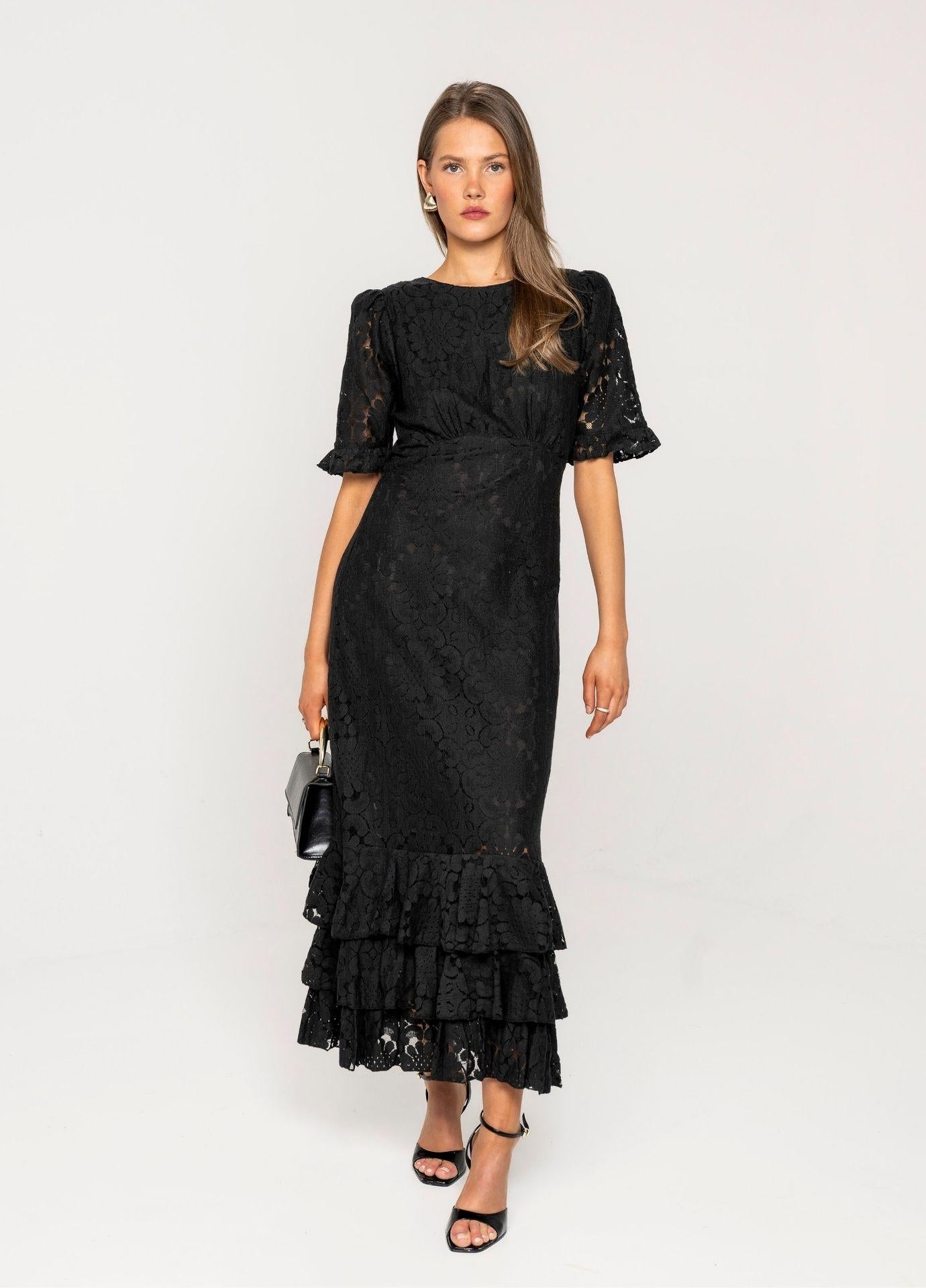 Odette Dress Noir Net in a full length front view, showing the gathered waist and the layered ruffle hem of the openwork black lace dress, with black strappy heels.